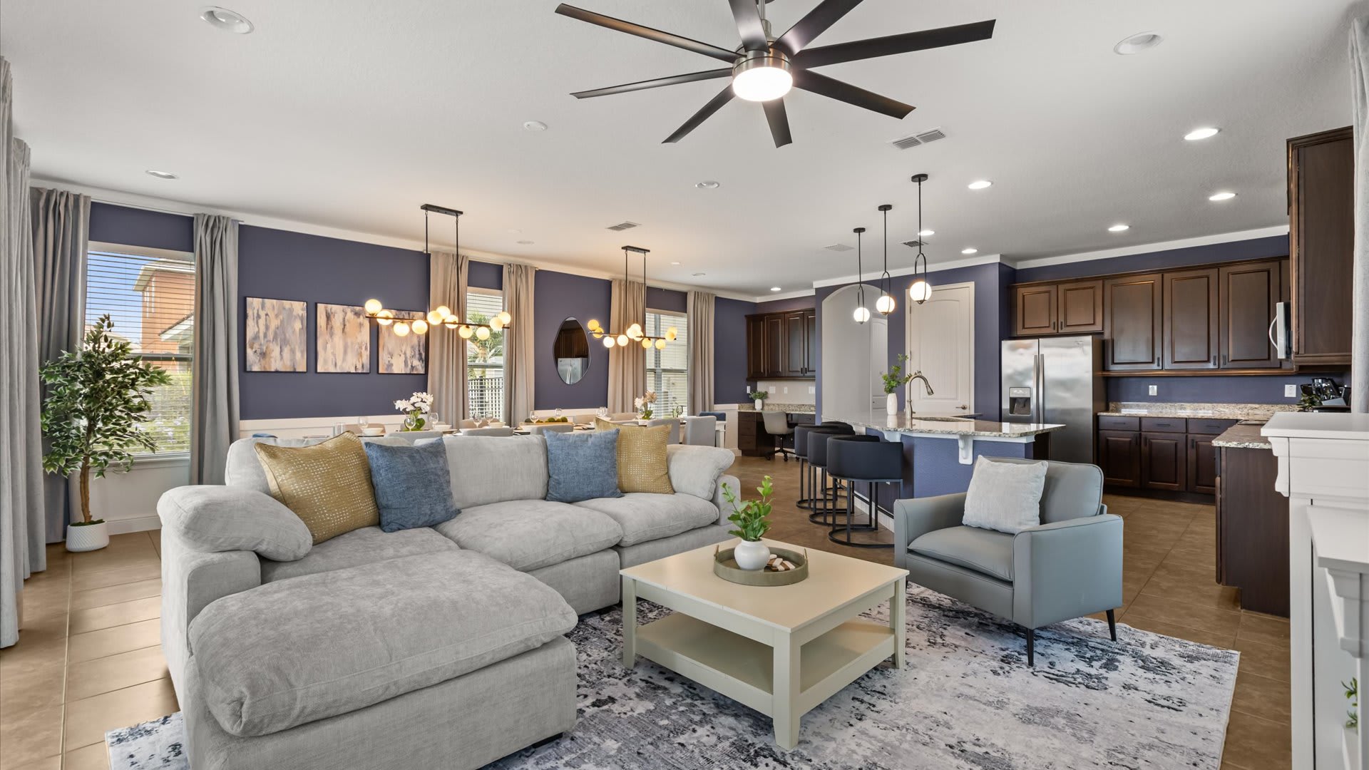 A spacious open-concept living room features a large light grey sectional sofa, a matching armchair, and a coffee table on a patterned area rug, with the kitchen visible in the background.