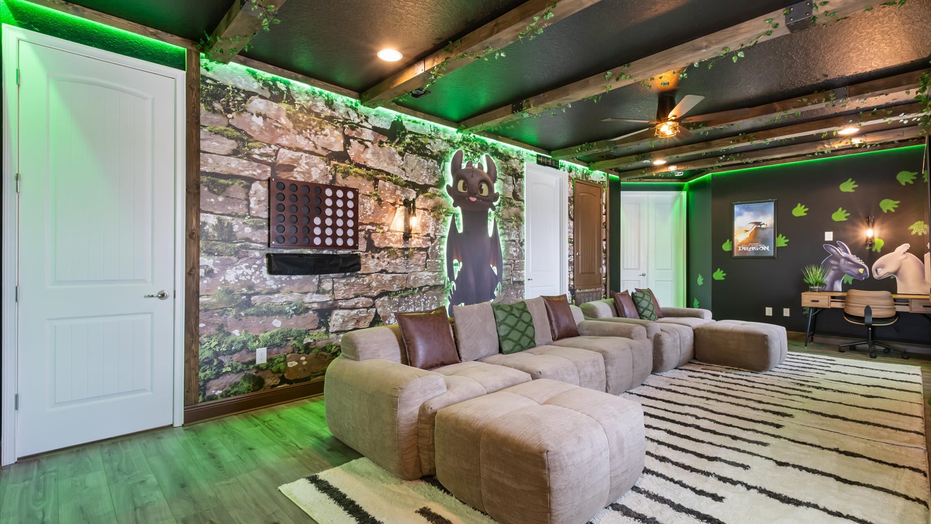 This "How to Train Your Dragon" themed media room showcases a large sectional sofa facing a stone-look accent wall with a Toothless cutout, illuminated by green LED lighting and featuring a small desk area.