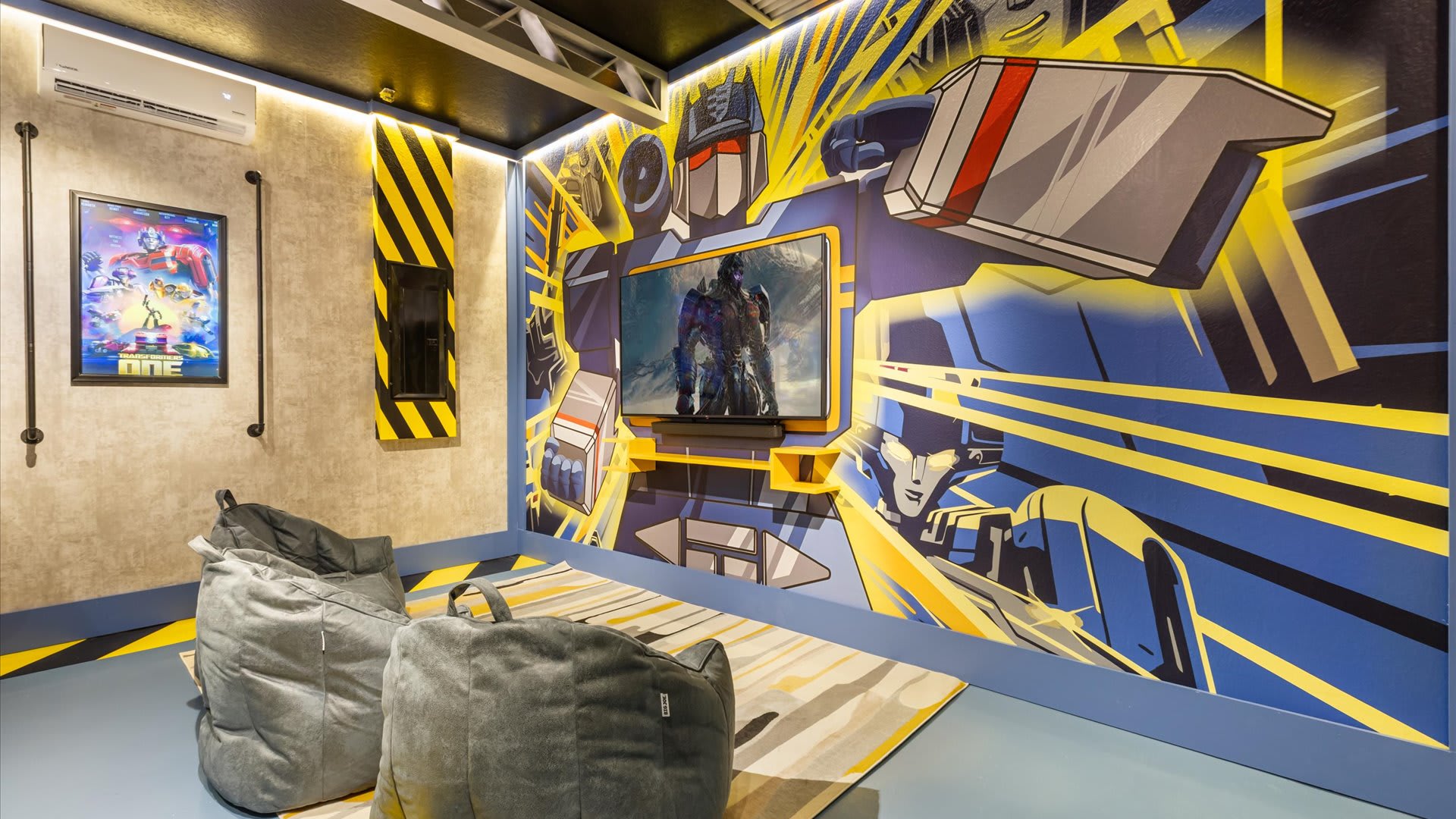 A dedicated media corner within the game room, featuring a large wall-mounted TV surrounded by a vibrant Transformers mural. Two grey bean bag chairs sit on a striped rug, providing comfortable seating for gaming or movie watching.