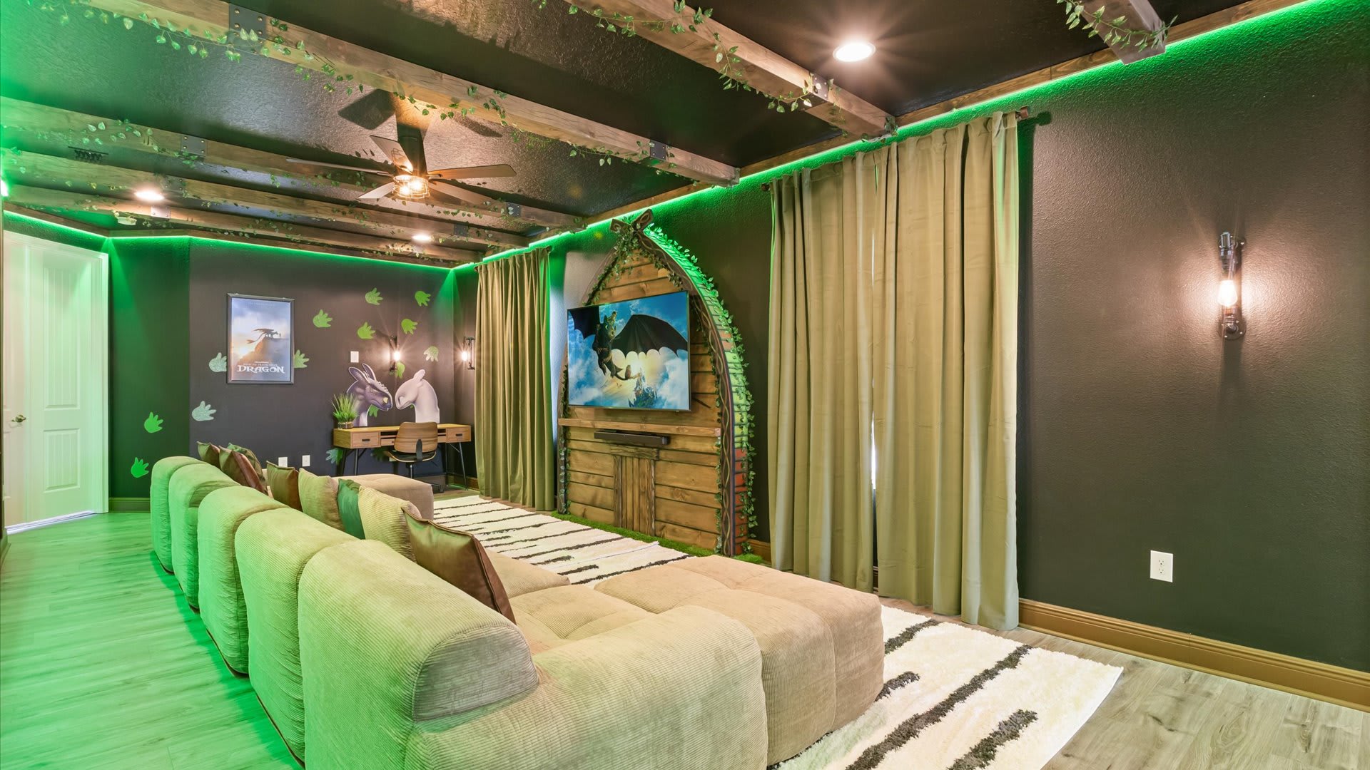 Themed media room with a large TV mounted in a rustic wooden frame, a long sectional sofa, and dark green walls with green LED lighting. Light-colored curtains frame the TV area, and the floor has a striped rug.
