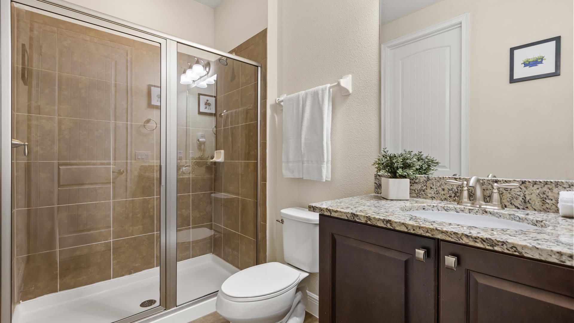 A clean bathroom with a dark wood vanity, granite countertop, and a glass-enclosed shower with brown tiled walls, featuring a white toilet and light-colored walls.