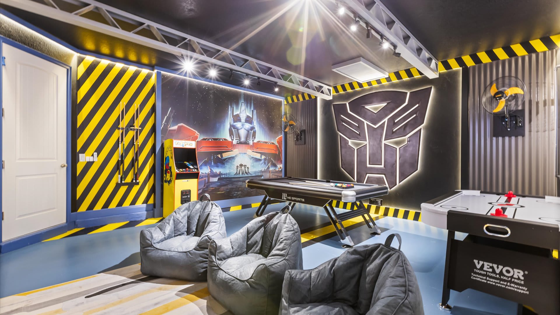 A vibrant Transformers-themed game room features a pool table, air hockey, and an arcade machine, complemented by three grey bean bag chairs and striking yellow and black striped walls.