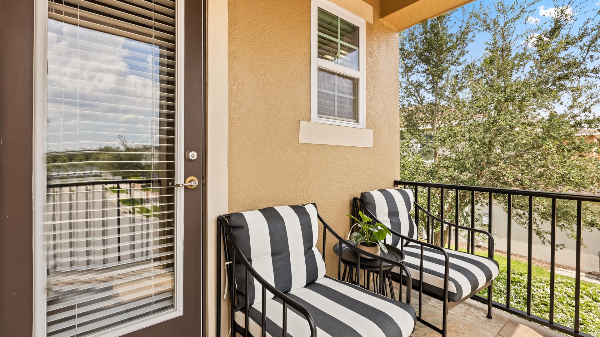 A small outdoor balcony with a black metal railing, furnished with two black and white striped chairs and a small side table, overlooking the property's exterior.