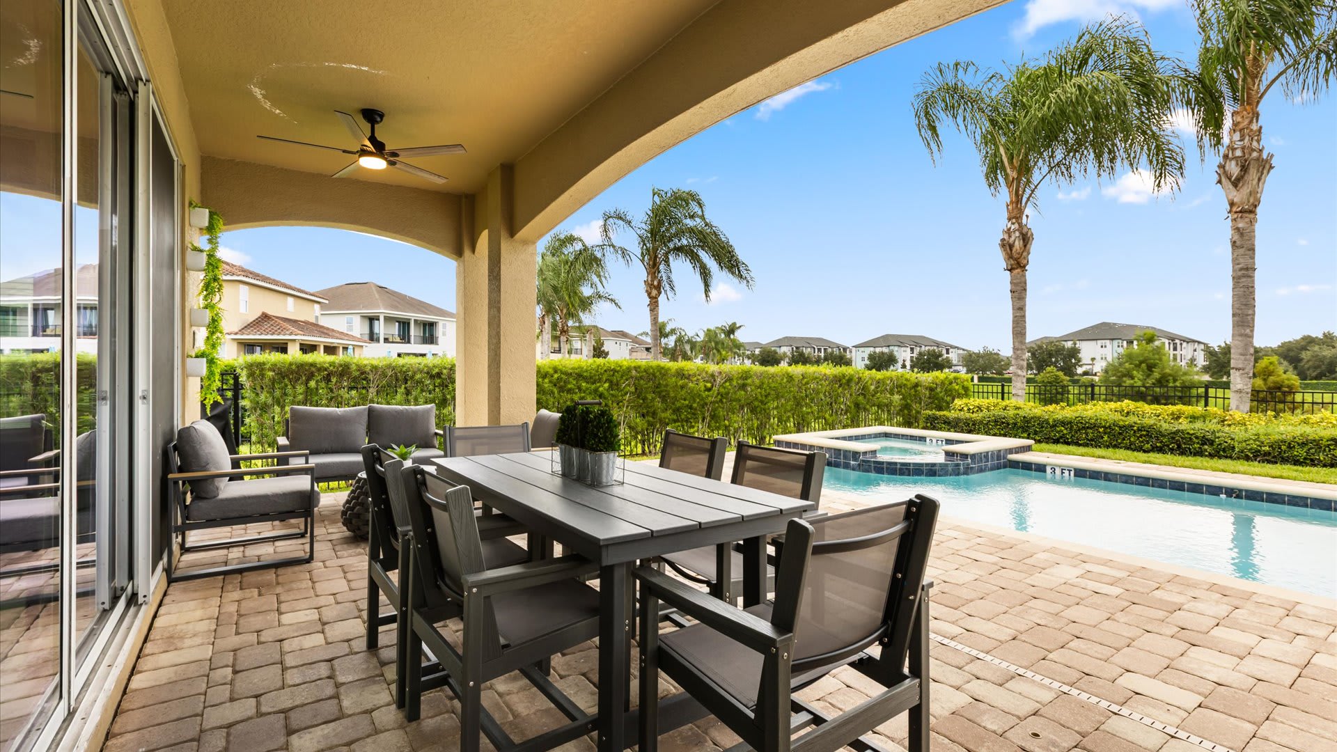 A covered outdoor patio features a dining table with six chairs and a separate lounge seating area, all on a paver patio overlooking the private pool and hot tub. A ceiling fan provides comfort.