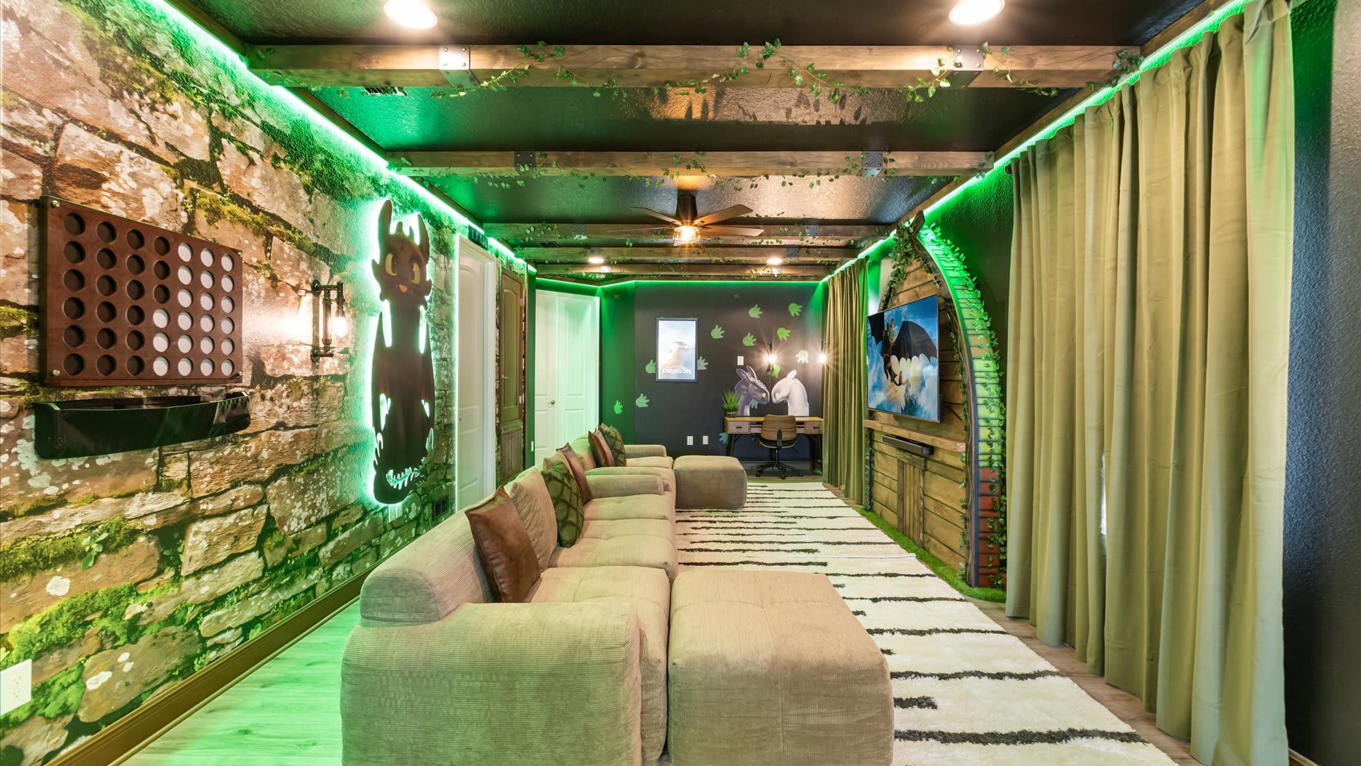 Wide view of the themed game room, showcasing the long sectional sofa, stone-look accent wall with a game board, and a desk area with dragon-themed decor. Green LED lighting illuminates the space, and the ceiling has decorative beams.