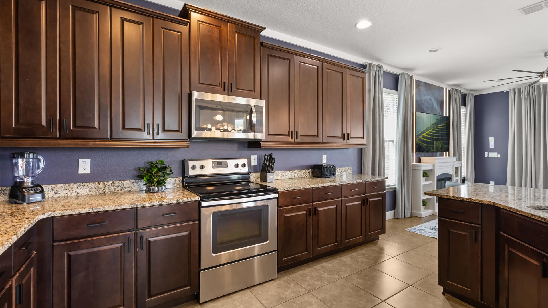 Modern kitchen with dark wood shaker cabinets, light granite countertops, stainless steel appliances including a range and microwave, and a dark purple accent wall. The floor is tiled in a light brown.