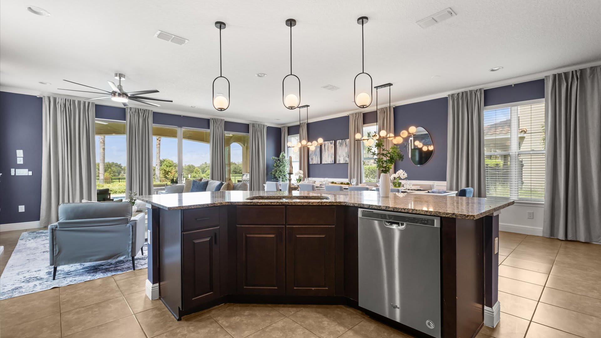 Spacious kitchen island with dark wood cabinetry and light granite countertops, featuring a stainless steel dishwasher and sink. Modern pendant lights hang above, and the open-concept space extends to a living area with large windows.