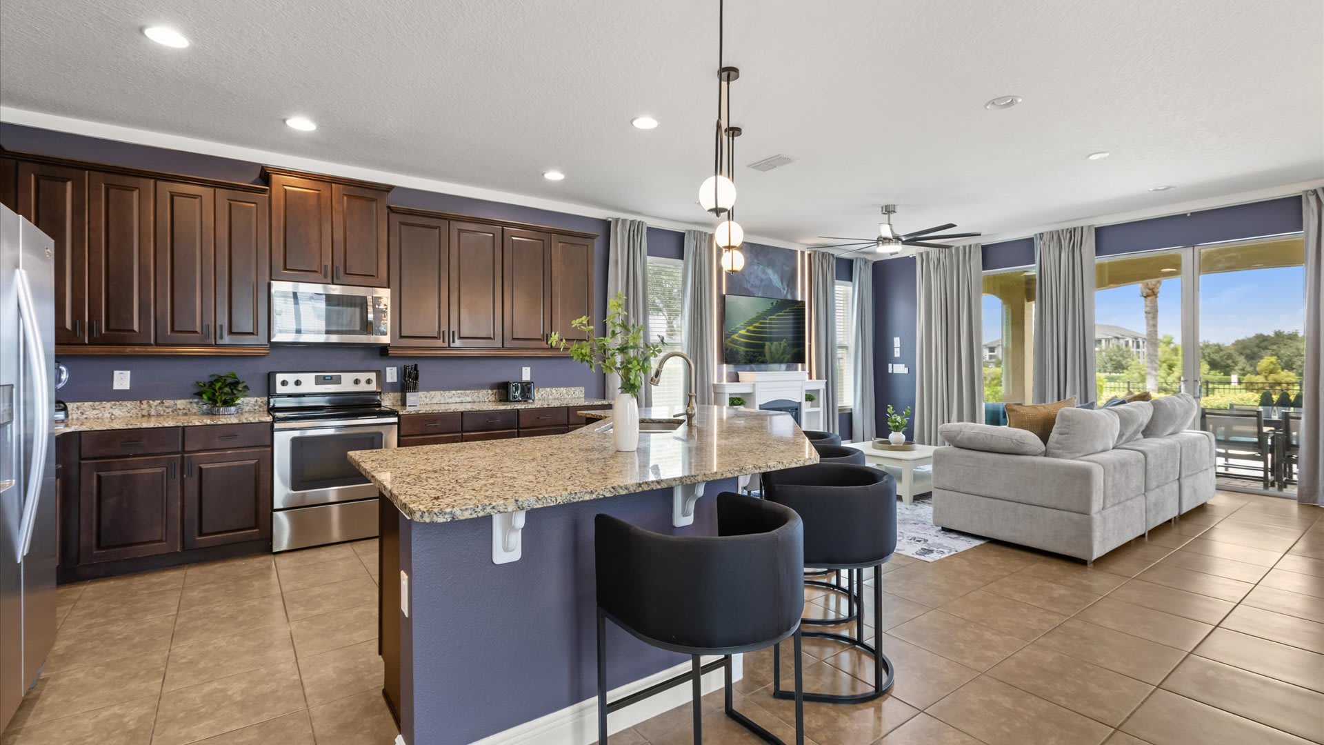 A spacious kitchen area showcases dark wood cabinets, granite countertops, and stainless steel appliances, centered around a large island with bar seating, flowing into the dining and living areas.