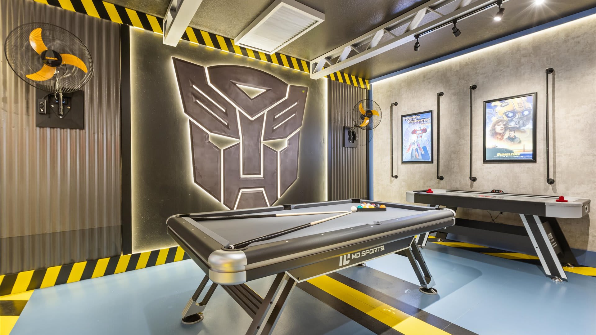 A vibrant game room, themed with a large illuminated Transformers logo, features a pool table and an air hockey table. Industrial-style fans and track lighting are mounted on the walls and ceiling.
