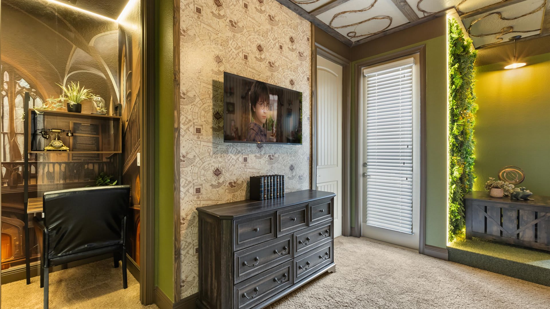 A section of the themed bedroom featuring a dark wood dresser with a wall-mounted TV, a small desk area with a chair, and a door with blinds providing natural light.