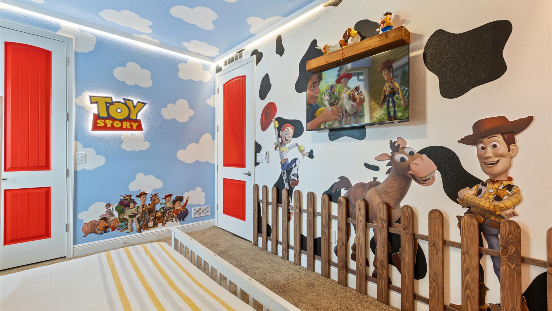 Another view of the Toy Story themed bedroom, showcasing a wall mural with Woody, Jessie, and other characters, a cow-print accent wall, and a wall-mounted TV above a wooden shelf.