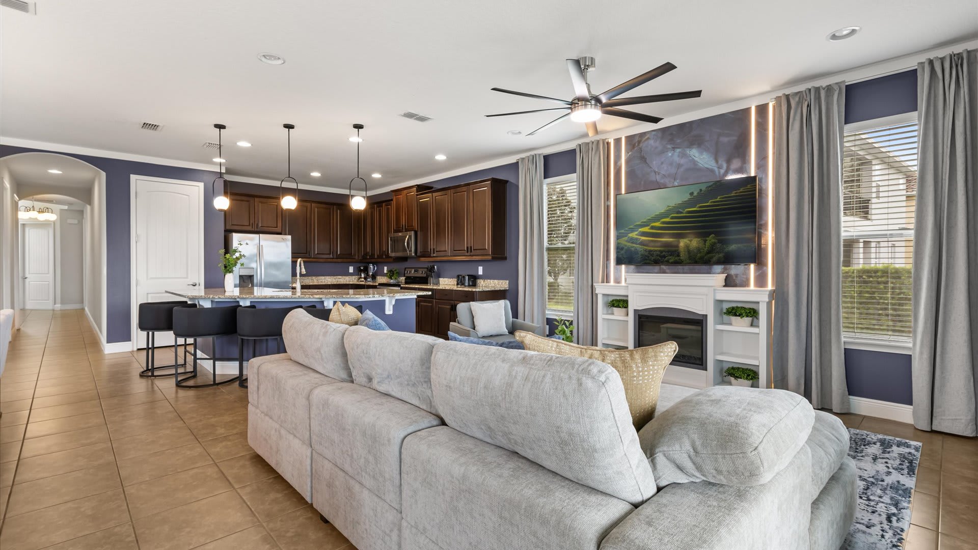 A comfortable living room with a large light grey sectional sofa faces a wall-mounted TV above a white fireplace, with the kitchen island and dining area visible in the open-concept space.