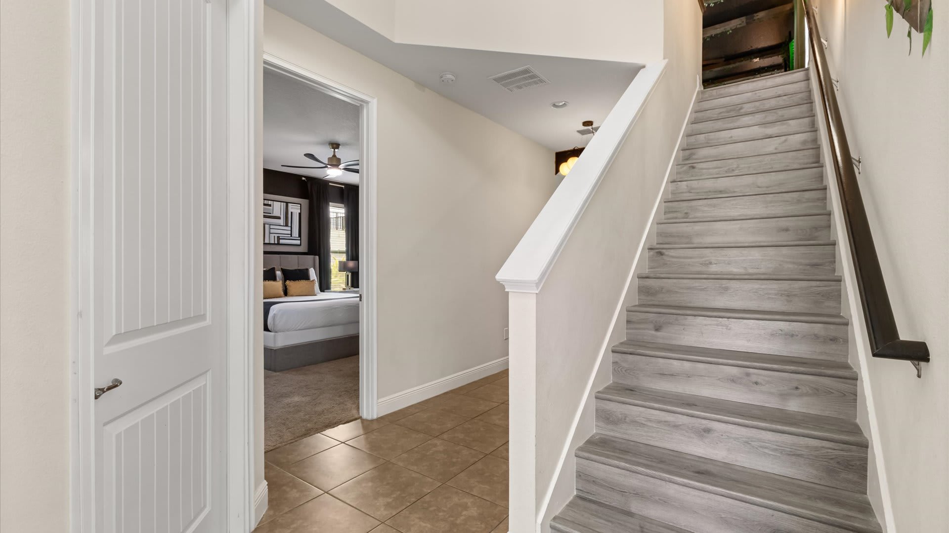 View of a staircase with light wood-look treads and white risers, leading upstairs. A white paneled door is visible to the left, opening into a bedroom, and the floor is light brown tile.