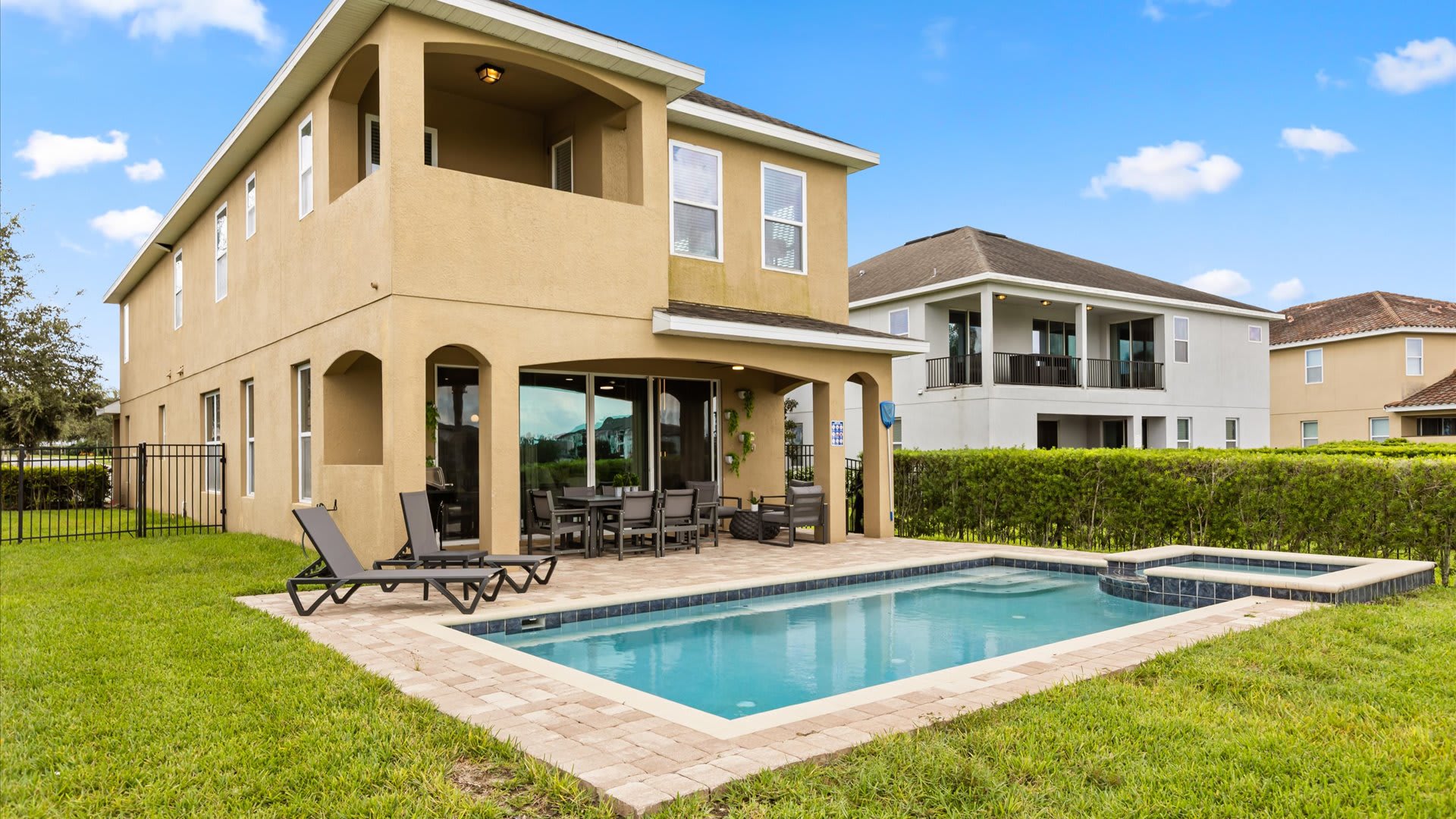 A wide shot of the two-story house's rear exterior, showcasing a private swimming pool and hot tub set within a manicured lawn. The covered patio features lounge chairs and an outdoor dining set, with a balcony on the second floor.