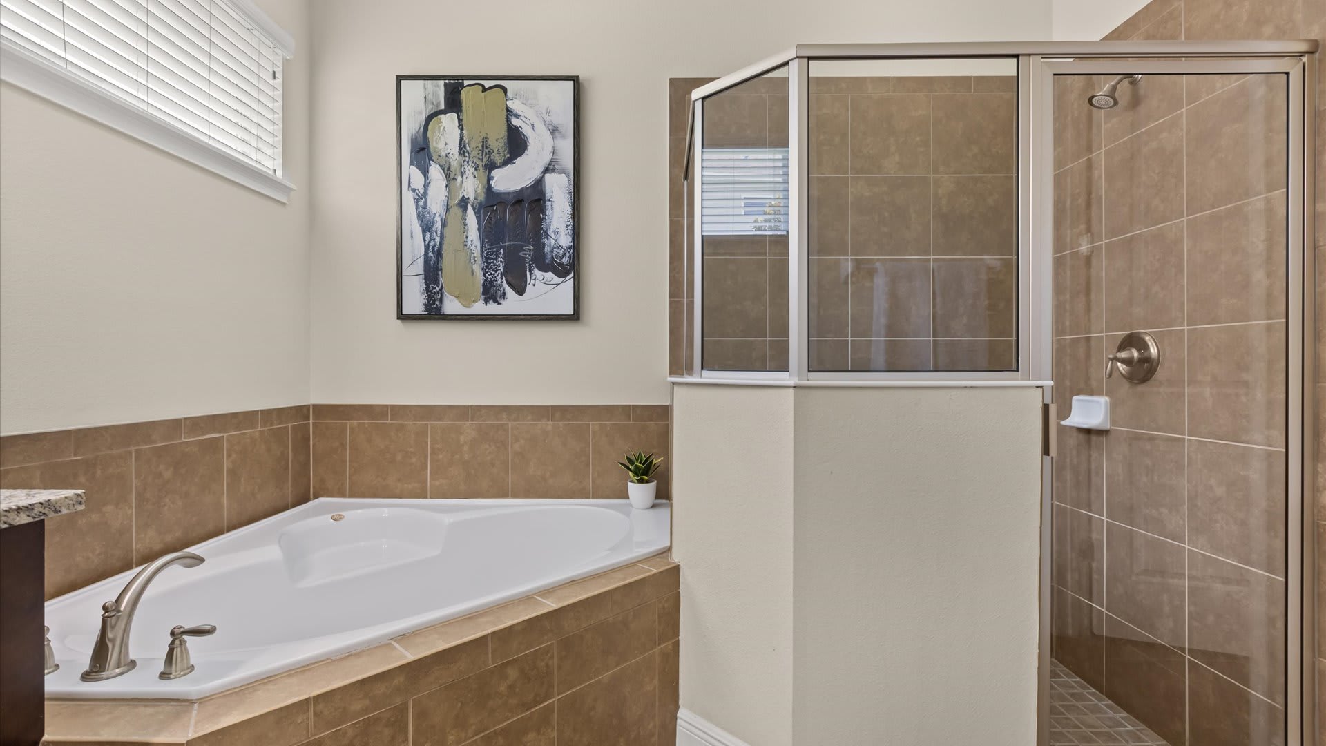 A spacious bathroom featuring a corner built-in jetted tub with a brushed nickel faucet, and a separate glass-enclosed shower with brown tiled walls. The walls are painted a light beige, complemented by a modern abstract painting.
