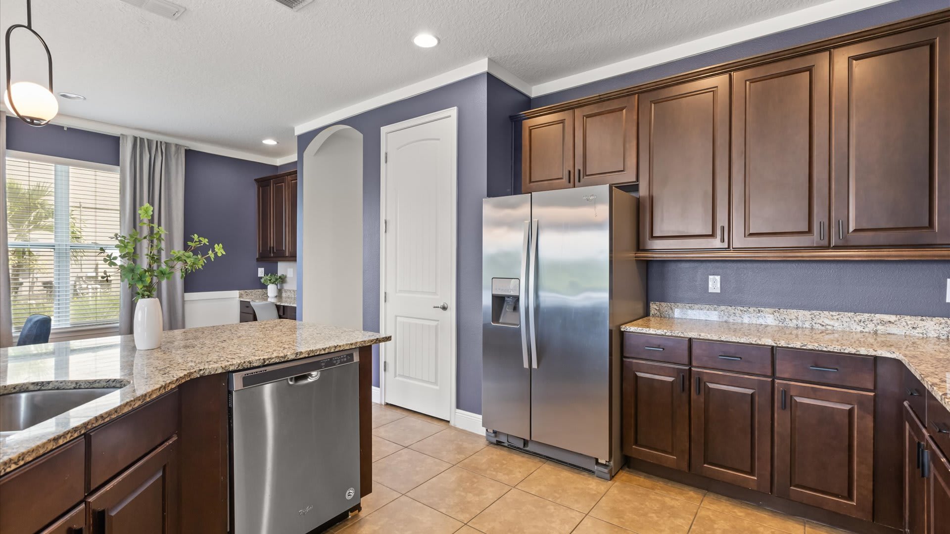 Kitchen area showing a stainless steel French door refrigerator and additional dark wood cabinets with granite countertops. A white arched doorway leads to another room, and the walls are painted a deep purple.