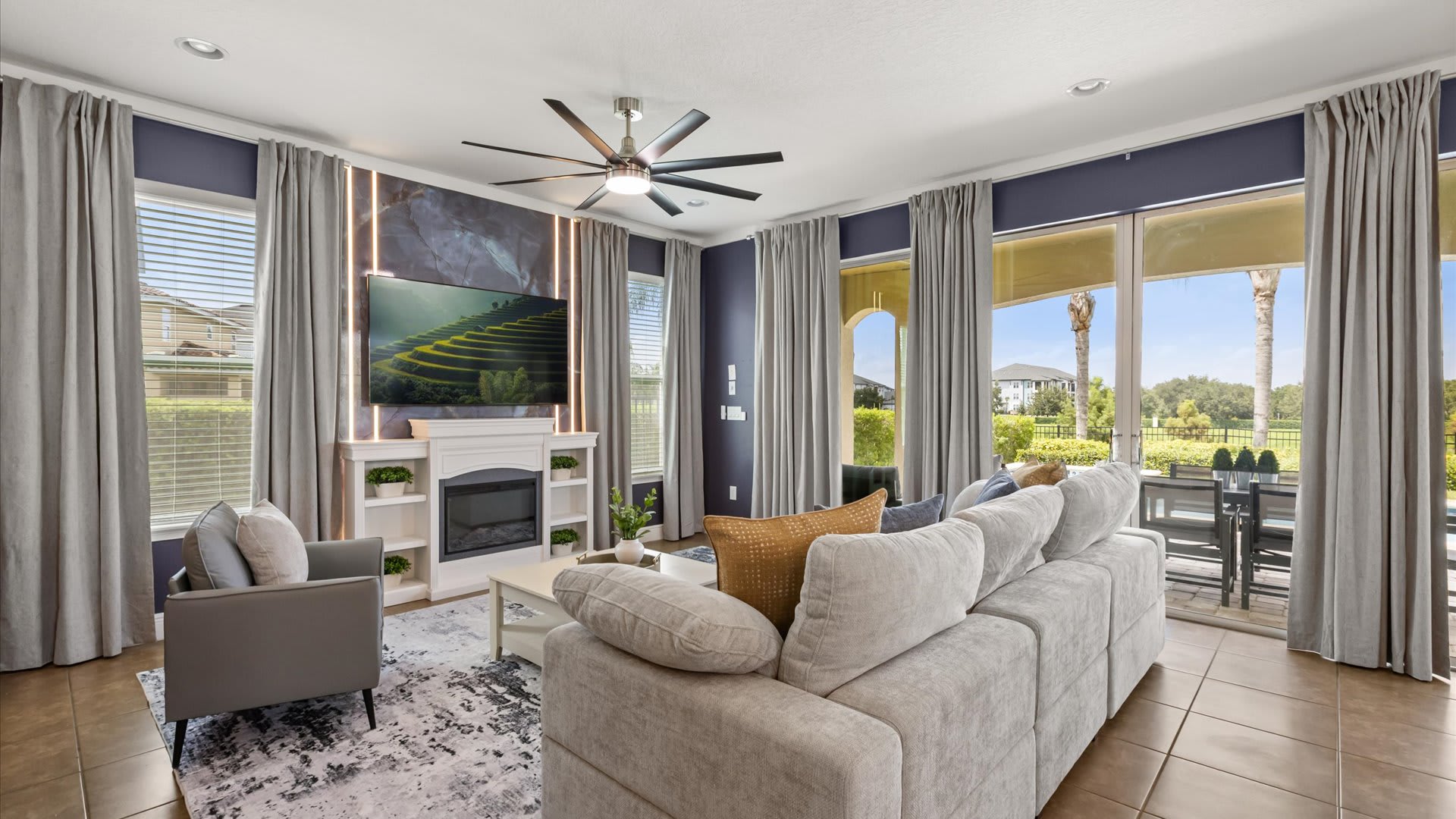 A modern living room features a large sectional sofa, a white fireplace with a mounted TV above, and floor-to-ceiling windows with dark blue walls and grey curtains. A ceiling fan with integrated lighting hangs overhead.