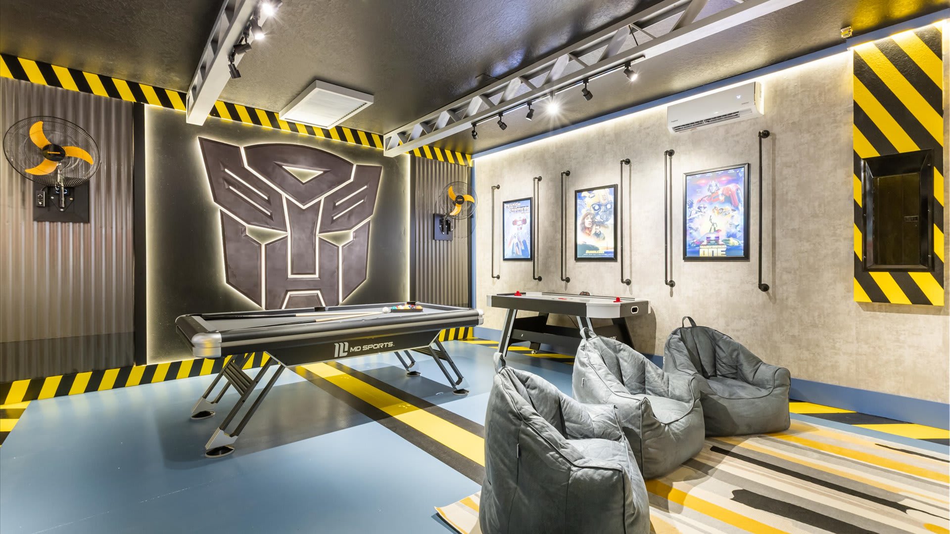 A vibrant game room with a Transformers theme, featuring a pool table, air hockey table, and grey bean bag chairs. The walls are decorated with corrugated metal panels, yellow and black striped accents, and a large illuminated Transformers logo.