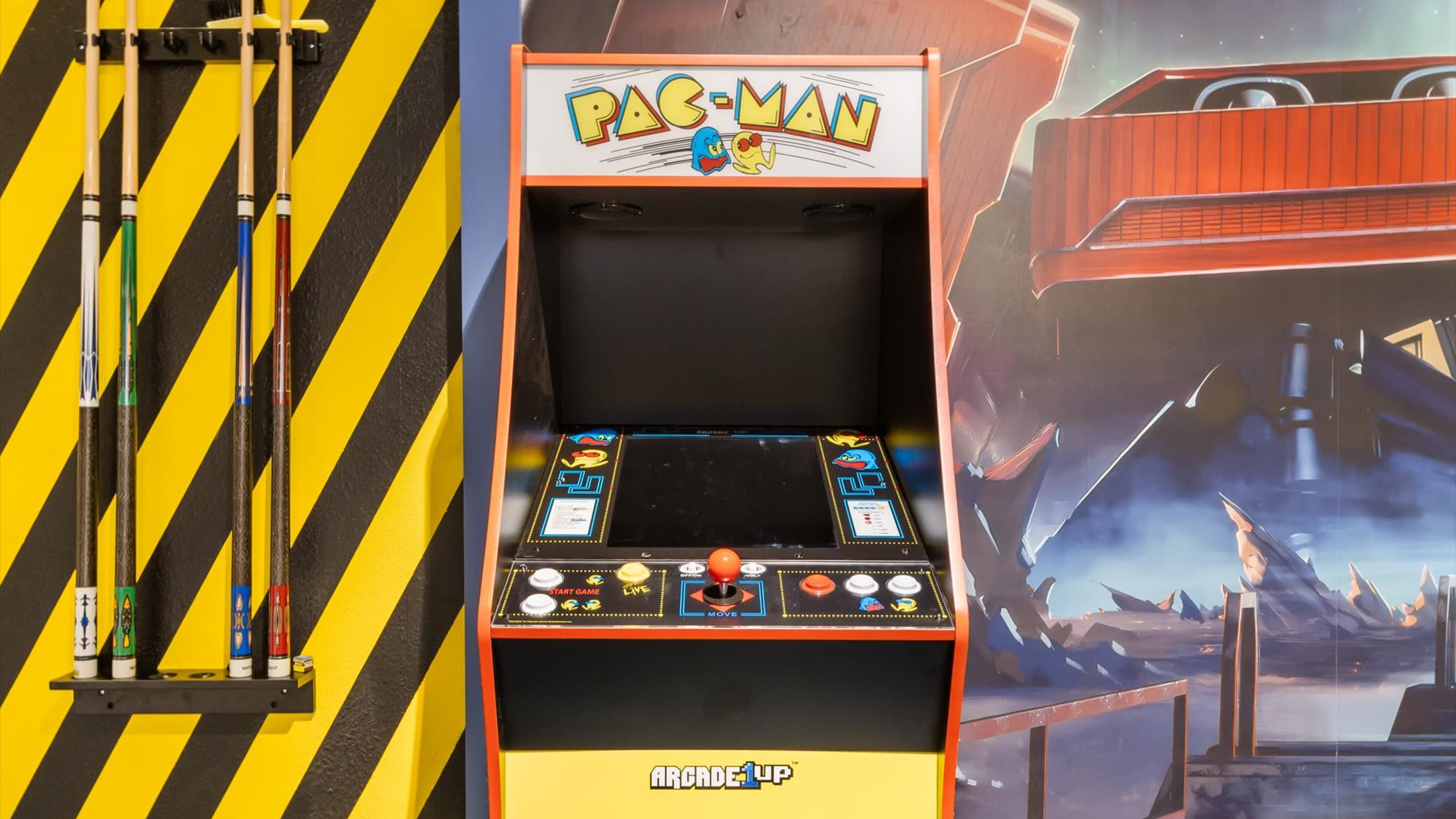 A close-up of the game room's arcade area, showcasing a classic Pac-Man arcade machine. The wall behind features a vibrant mural and yellow and black striped accents, with pool cues neatly hung on the wall.