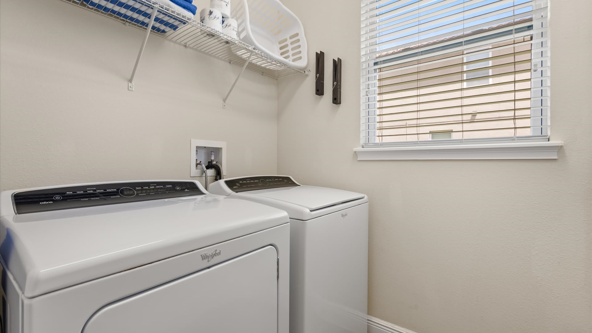 A bright and functional laundry room featuring a white front-loading Whirlpool washer and dryer set, a wire shelf with laundry baskets and towels, and a window with white blinds providing natural light. The walls are painted a light neutral color.