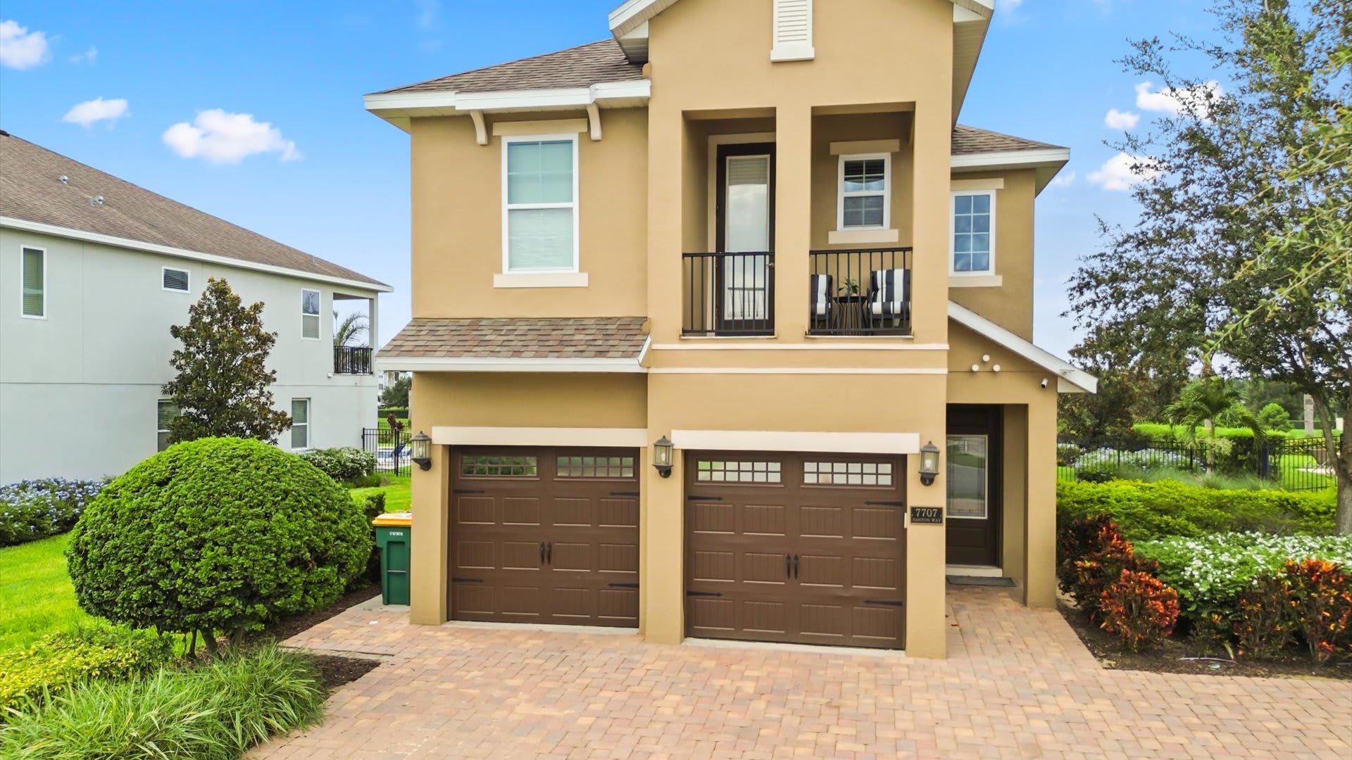 The front exterior of a two-story, tan stucco house with two brown garage doors and a small balcony on the second floor. A paved driveway leads to the garage, complemented by lush green landscaping and manicured bushes.