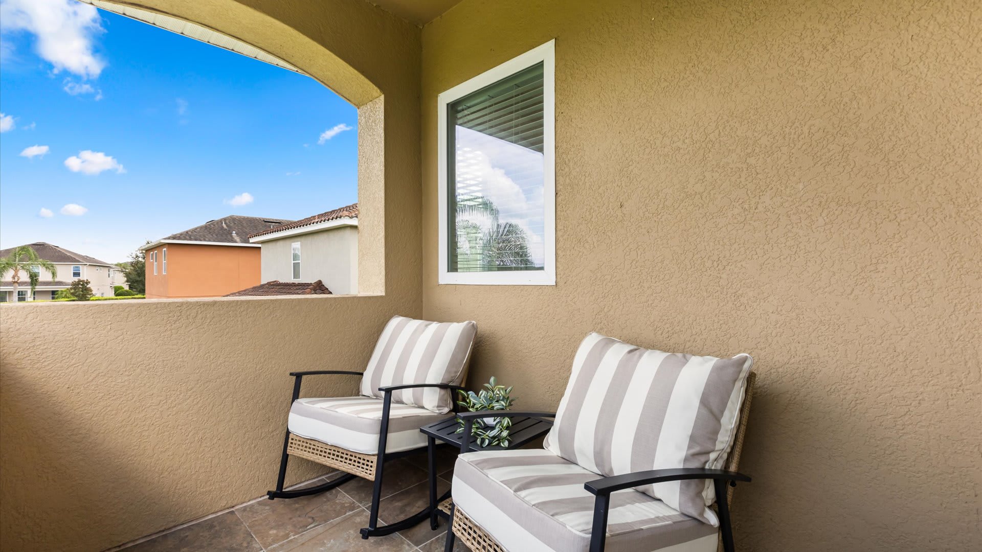 A cozy covered balcony featuring two striped rocking chairs with cushions and a small black side table, offering a view of the neighborhood.