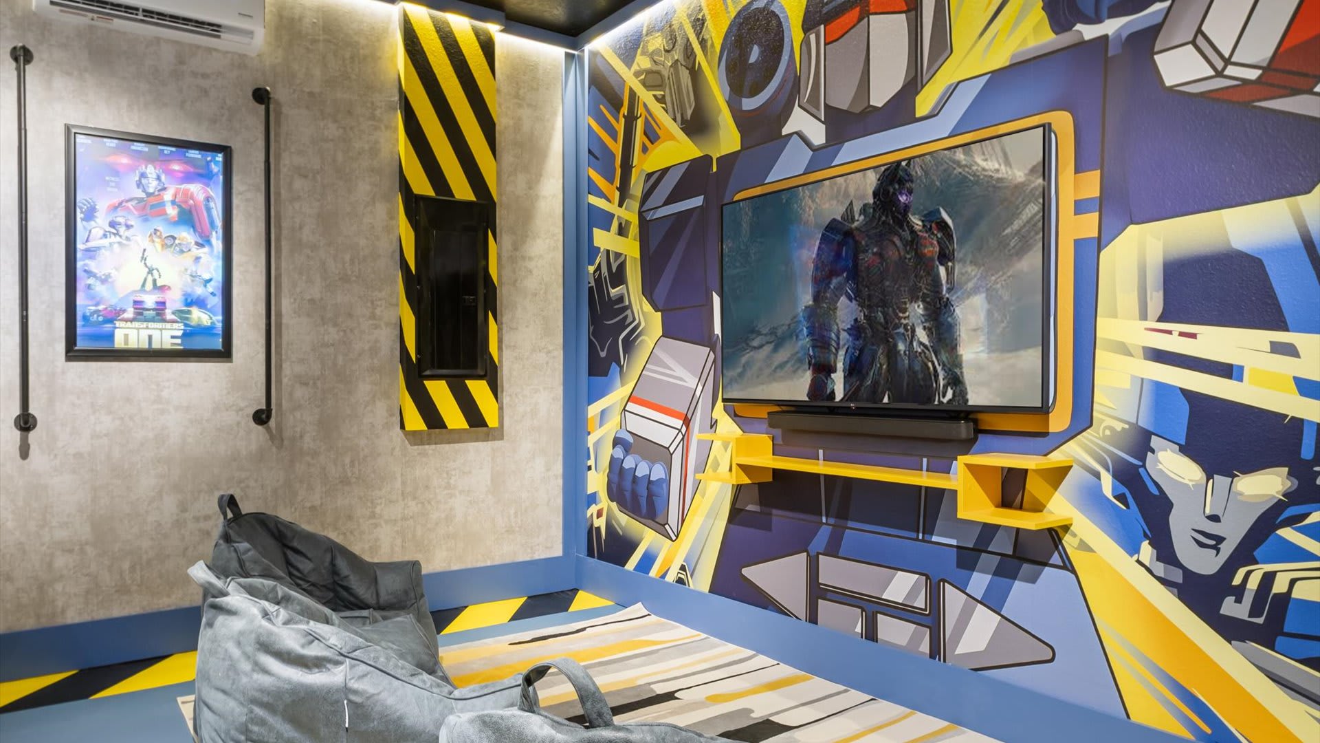 A cozy Transformers-themed media corner features a large wall-mounted TV with a soundbar, a grey bean bag chair, and a vibrant mural depicting Transformers characters on the walls.