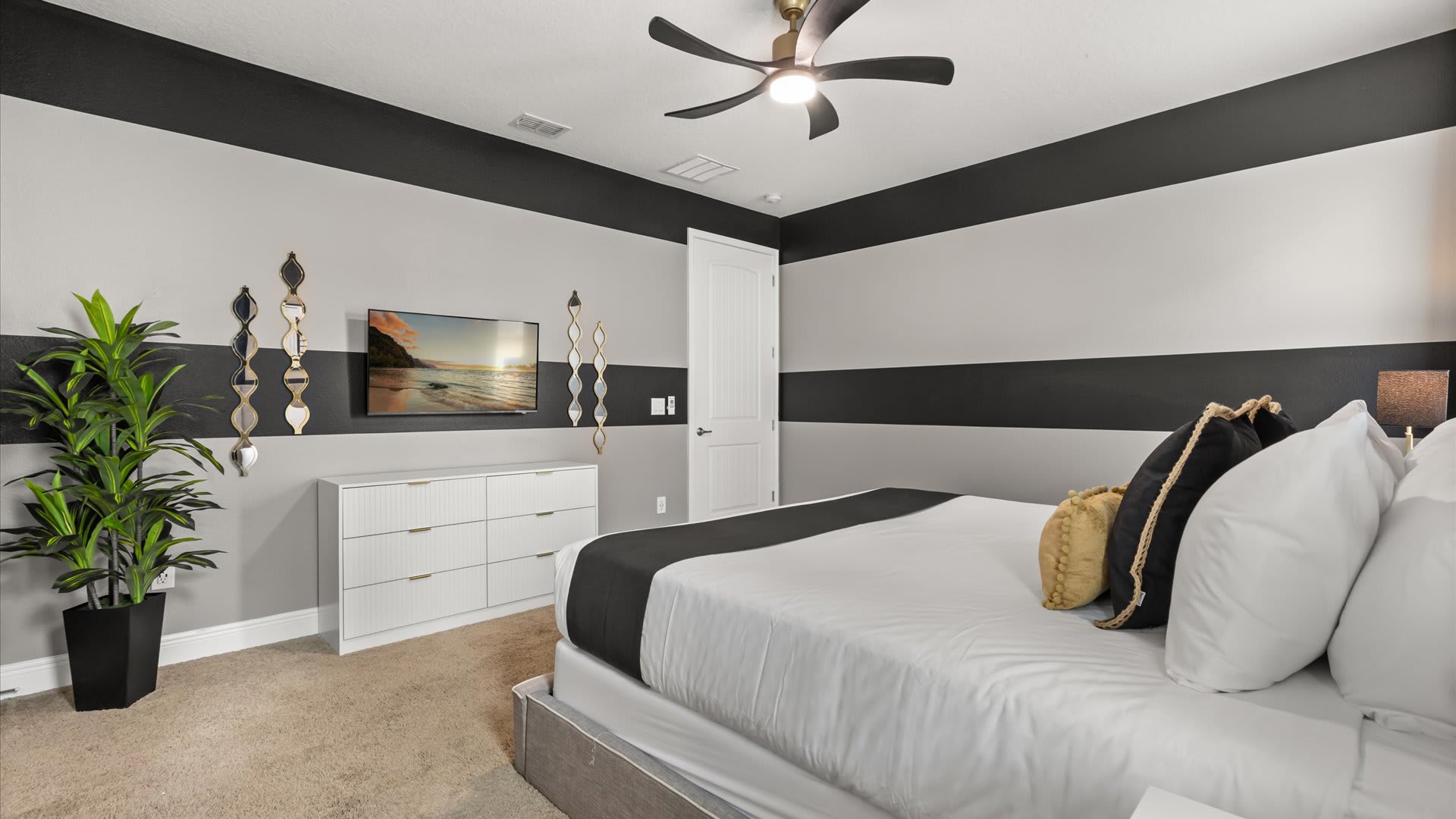 Another angle of the stylish bedroom, showing a wall-mounted TV above a white dresser, decorative wall mirrors, and a large potted plant in the corner.
