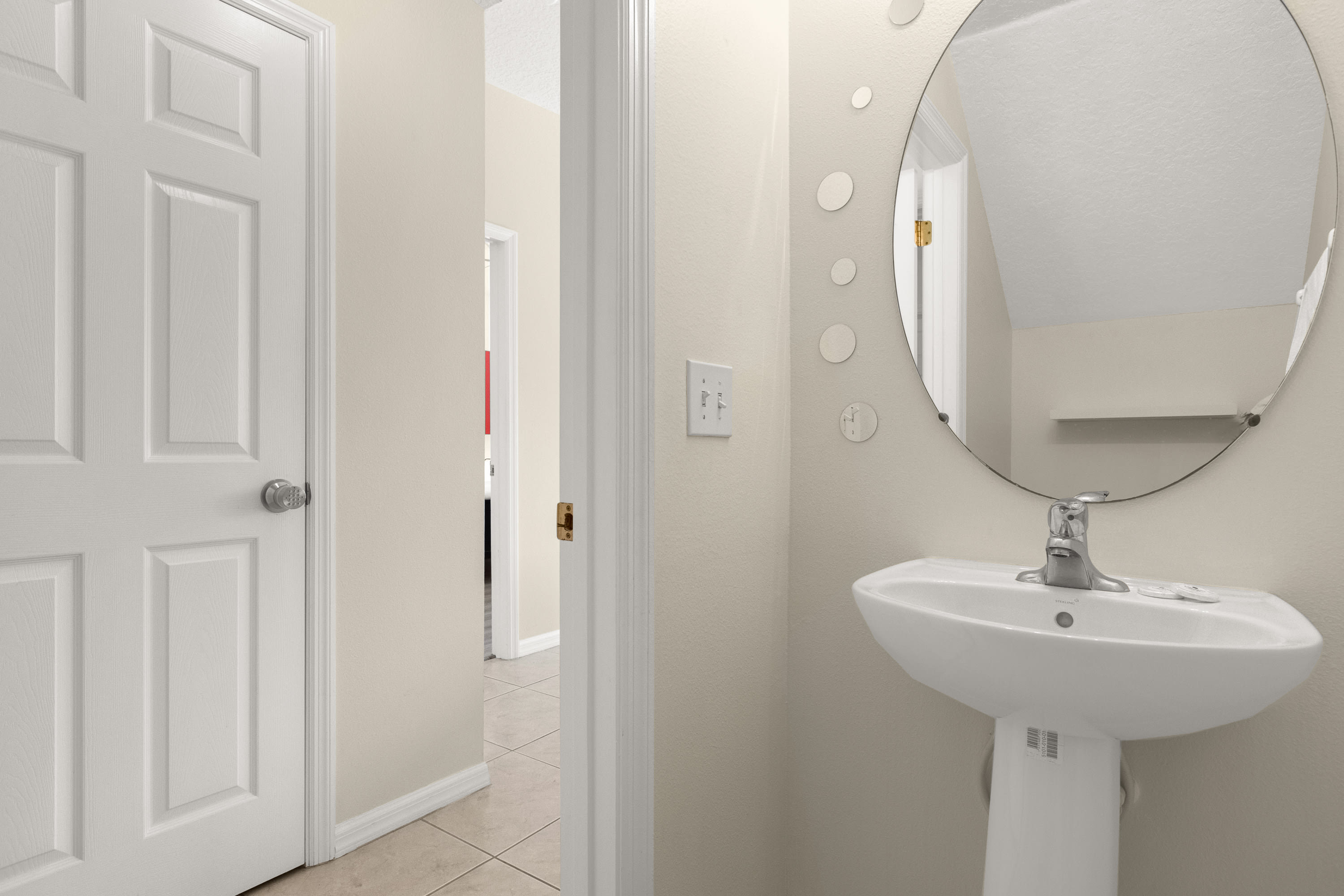 A compact half-bathroom with a white pedestal sink and a round mirror adorned with decorative circular accents, located near a white door and a hallway entrance.