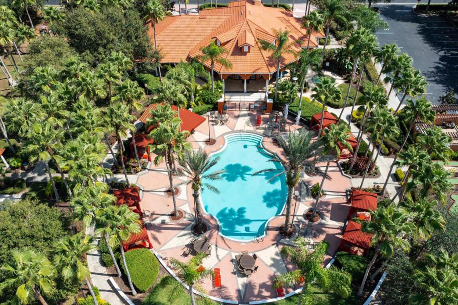 An aerial view showcases a large, irregularly shaped communal swimming pool surrounded by palm trees and red-roofed cabanas. The pool area is part of a resort complex with a central building, lush landscaping, and ample seating.