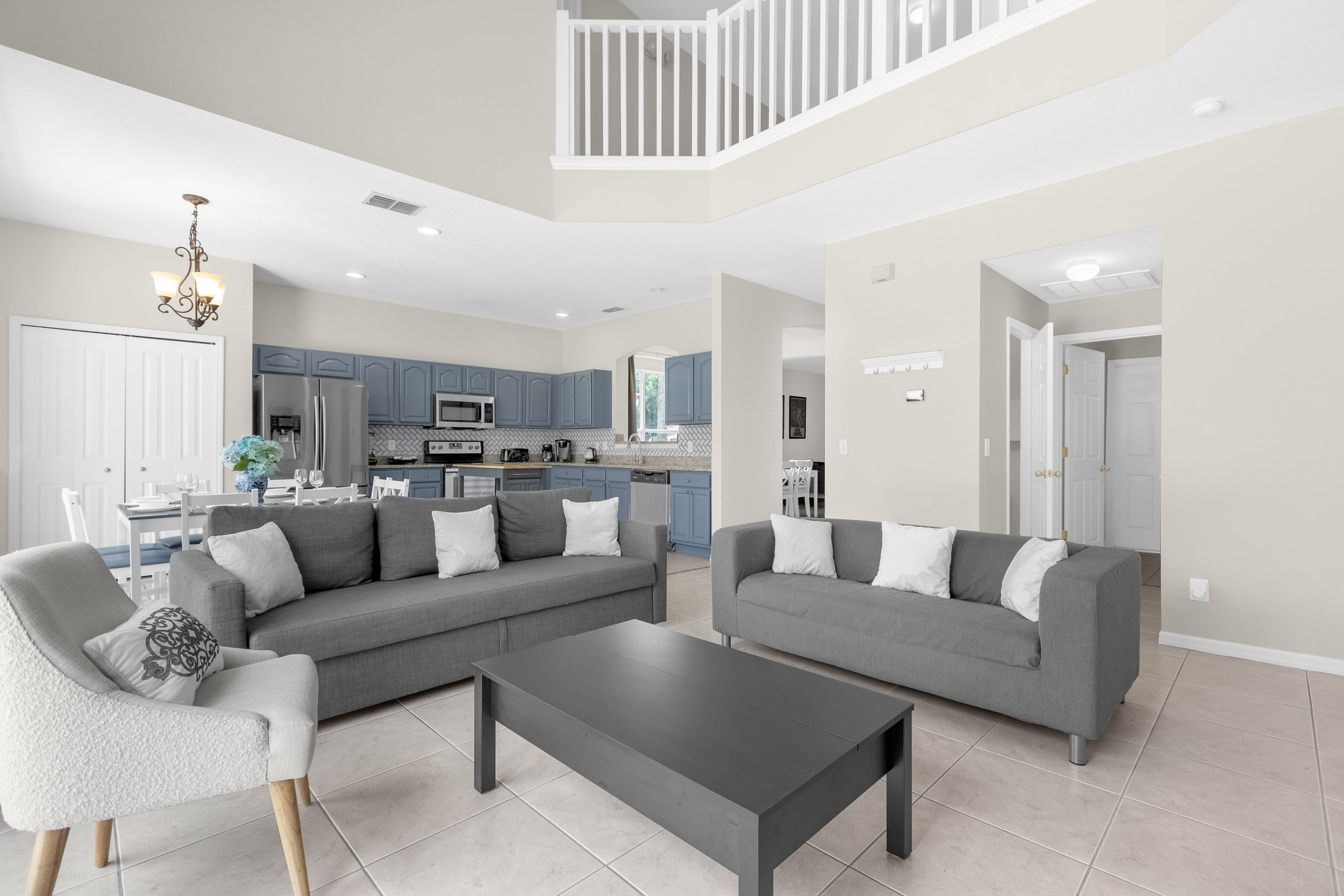 An open-concept living space features two gray sofas with white throw pillows, a dark gray coffee table, and a light armchair, all on light tile flooring. The area flows into a kitchen with blue cabinets and stainless steel appliances, and a dining area with a white table.