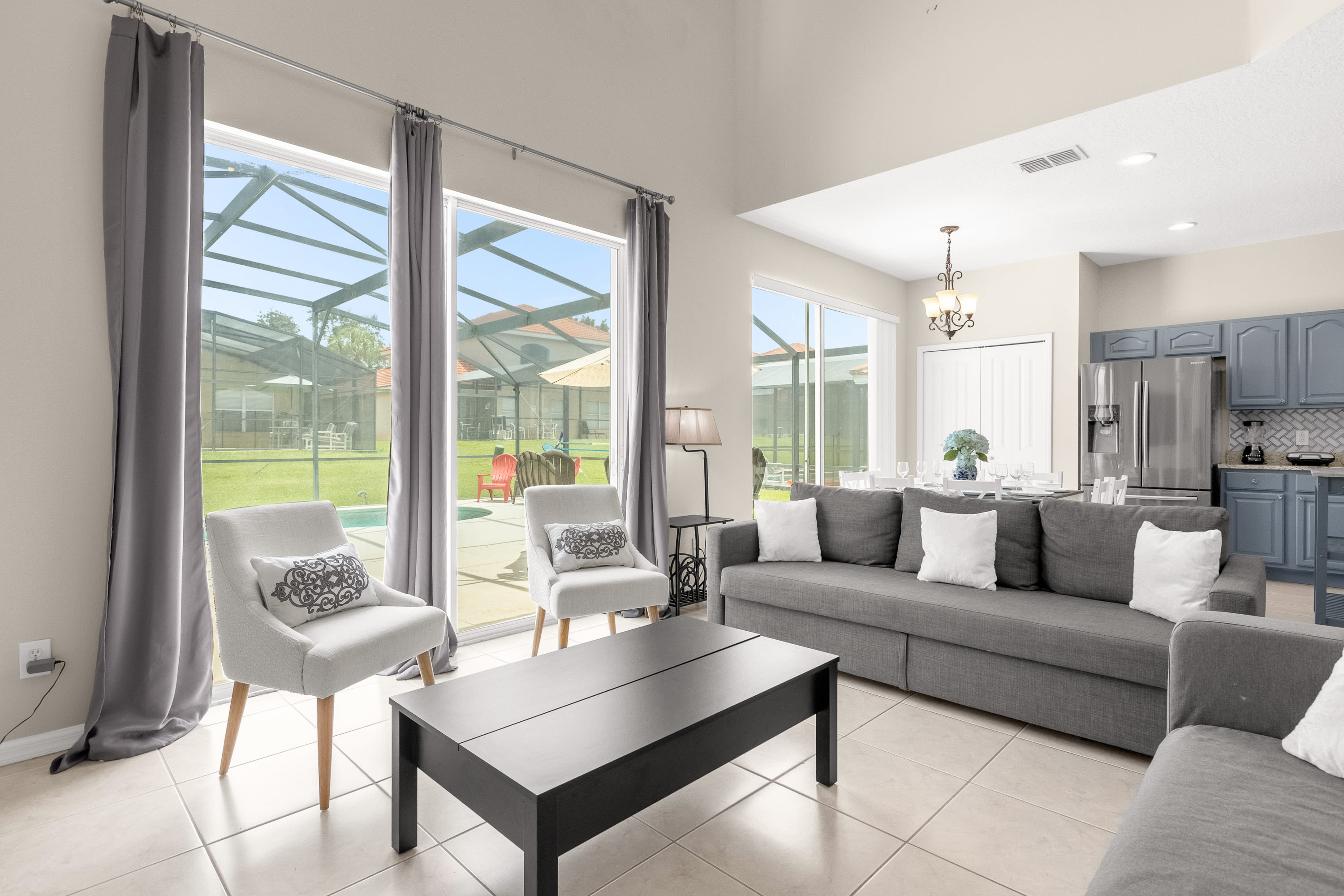 Bright living room with light tile flooring, a gray sectional sofa, two gray armchairs, and a black coffee table, featuring tall windows with gray curtains overlooking a private pool and backyard.