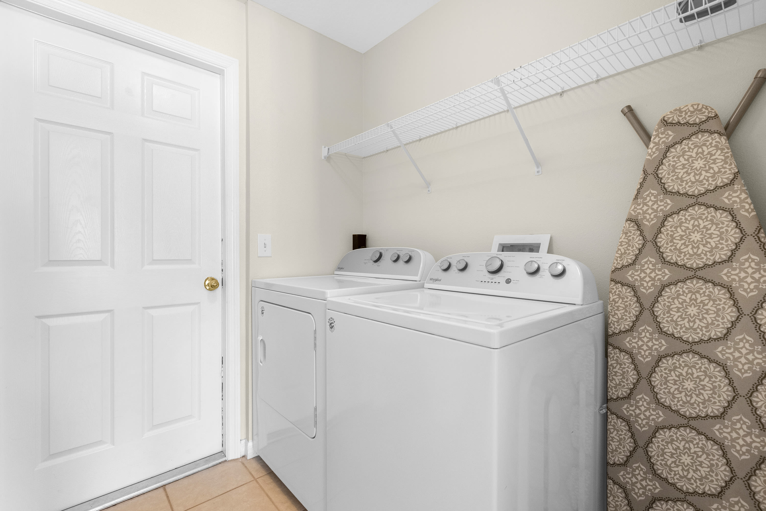 A functional laundry room equipped with a white top-loading washing machine and a matching dryer, featuring wire shelving above and an ironing board leaning against the wall.