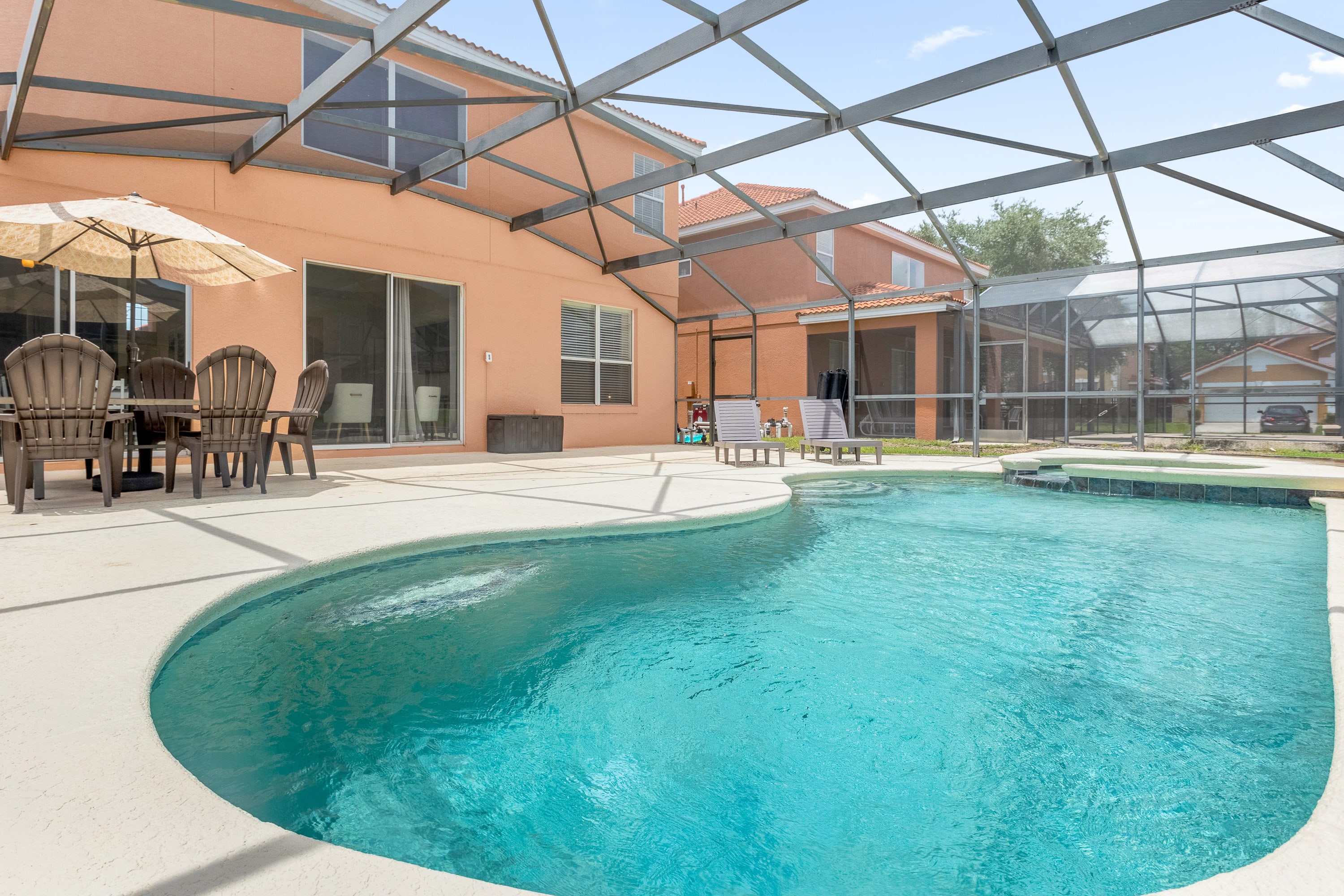 A wide view of the screened-in private pool area shows a large, freeform pool with a waterfall feature, surrounded by a spacious concrete deck. Two dark lounge chairs are positioned near the pool, with the peach-colored house exterior and sliding glass doors visible in the background.