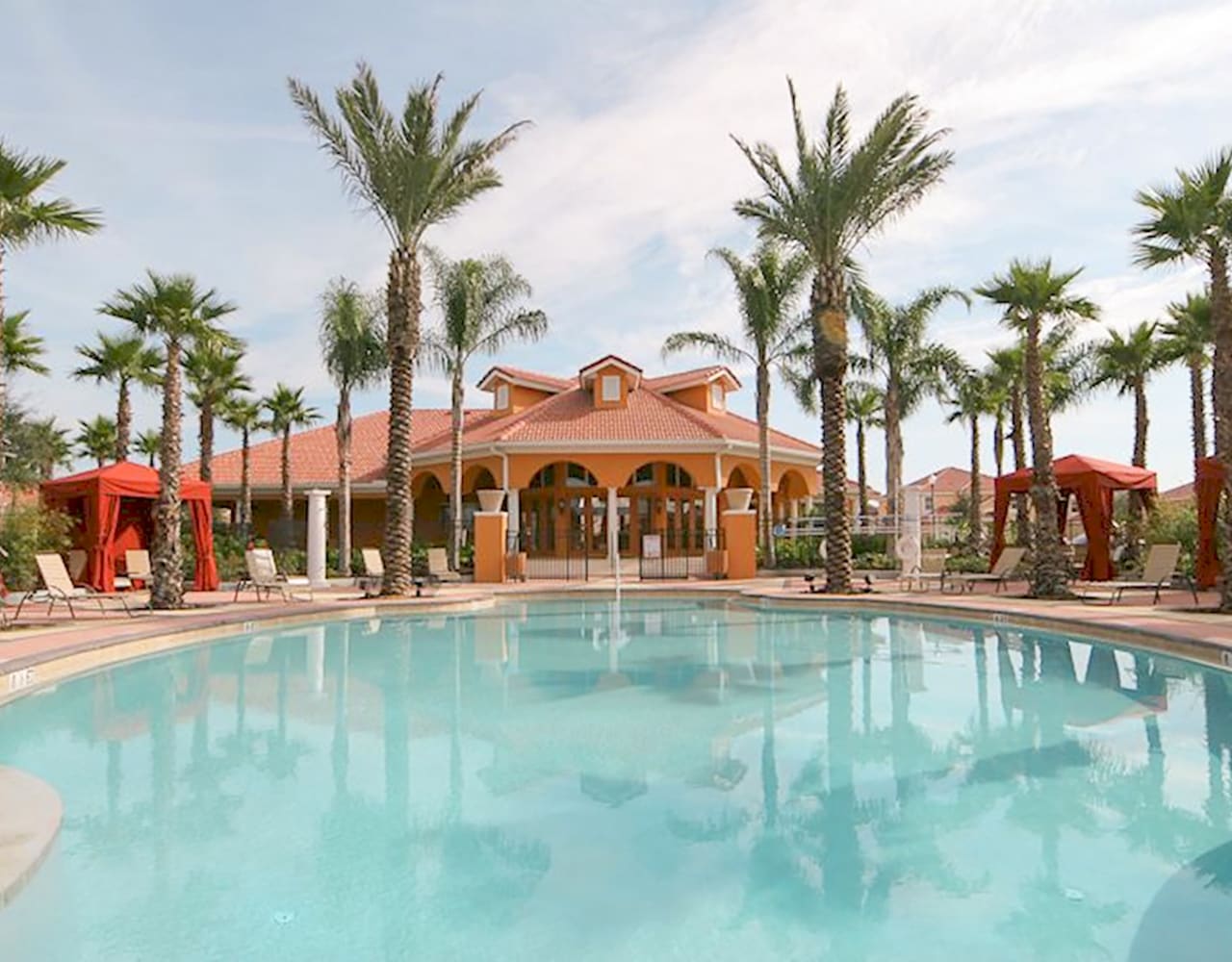 A large, inviting communal swimming pool with clear blue water, surrounded by tall palm trees and red cabanas. In the background, a large, orange-tiled clubhouse with arched windows and a terracotta roof stands under a bright sky.