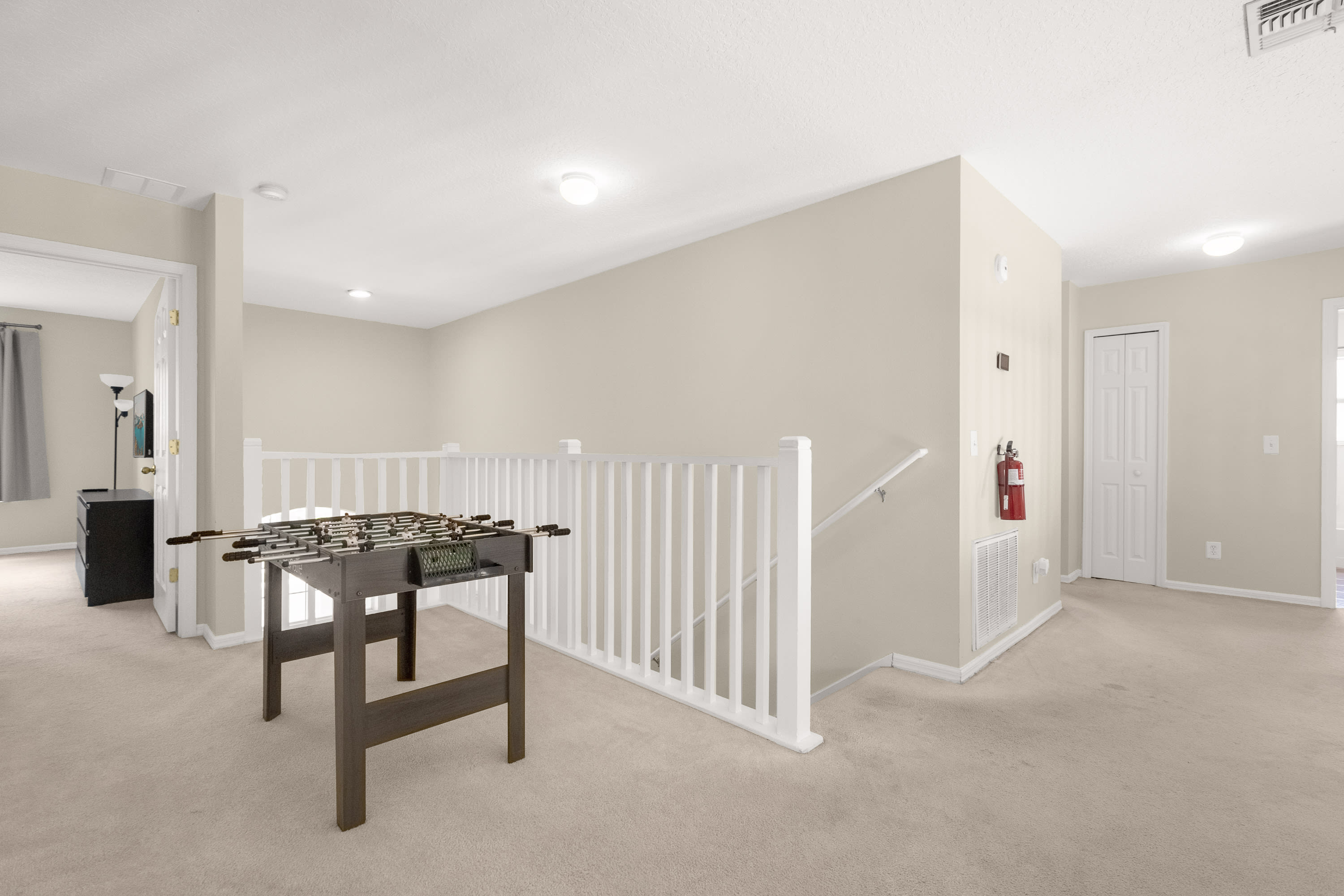 An upstairs loft area features a wooden foosball table on a light beige carpet, surrounded by white railings overlooking the floor below. A fire extinguisher is mounted on a light beige wall, and white doors lead to other rooms.