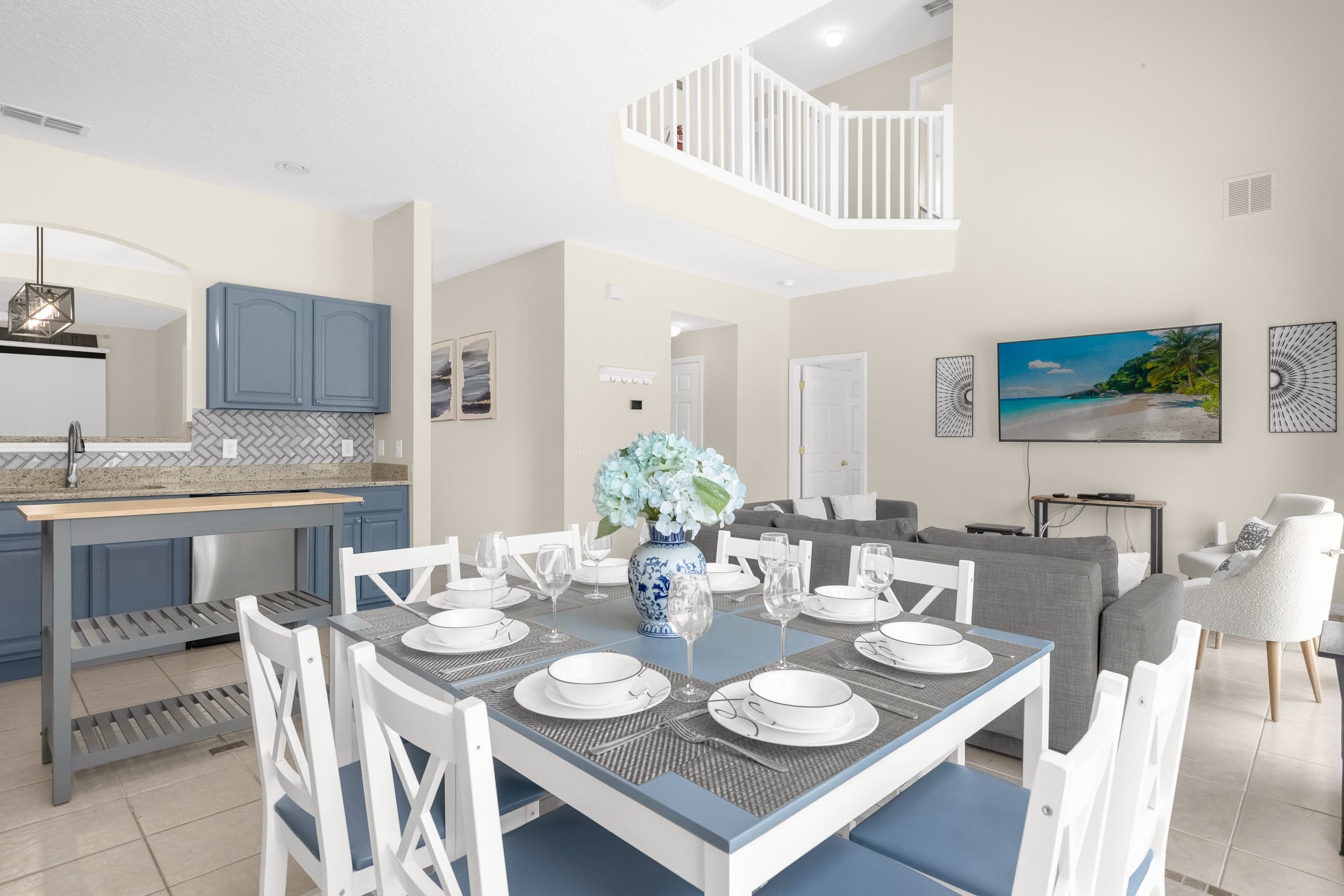 Open-concept dining area with a light blue and white dining table set for eight, featuring a blue floral centerpiece, adjacent to a kitchen with blue cabinets and stainless steel appliances, and a view of a staircase.