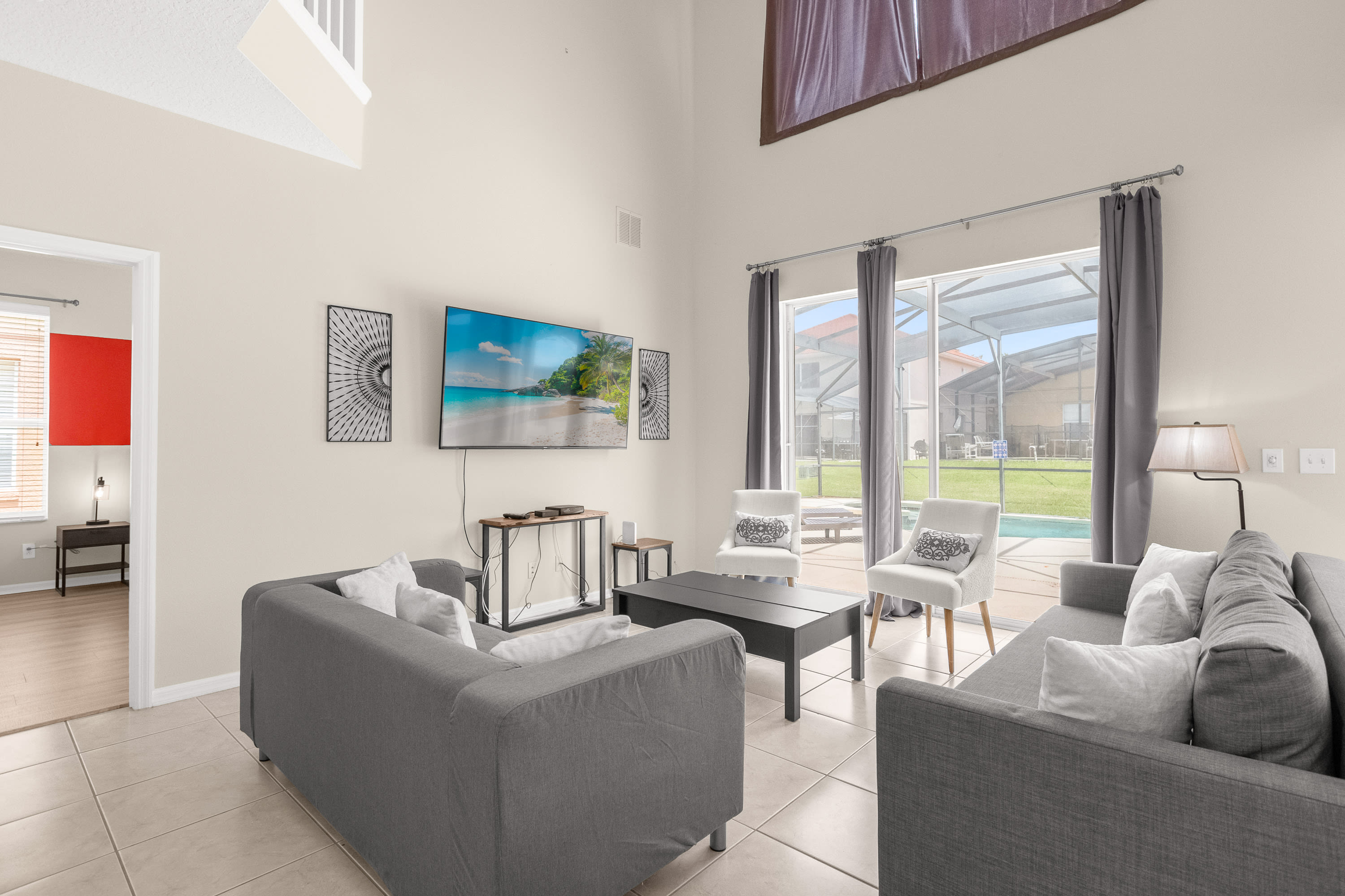 A bright living room features two gray sofas facing a wall-mounted flat-screen TV, with a dark coffee table in the center. Two white armchairs with patterned pillows are positioned near large sliding glass doors that open to the screened-in pool area, allowing natural light to fill the space.