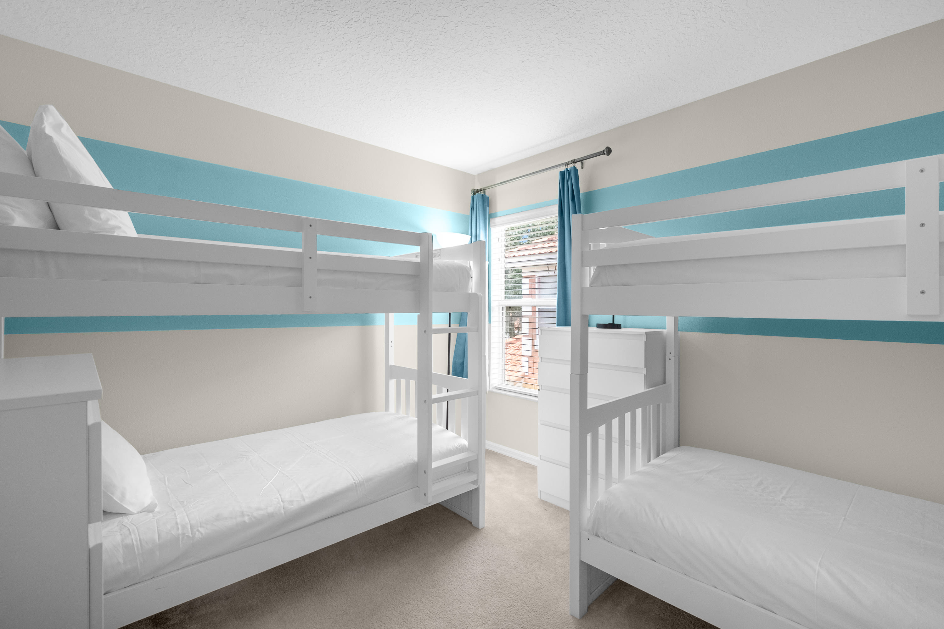 A children's bedroom featuring two white bunk beds with white bedding, set against a wall with horizontal blue and beige stripes, and a window with blinds.