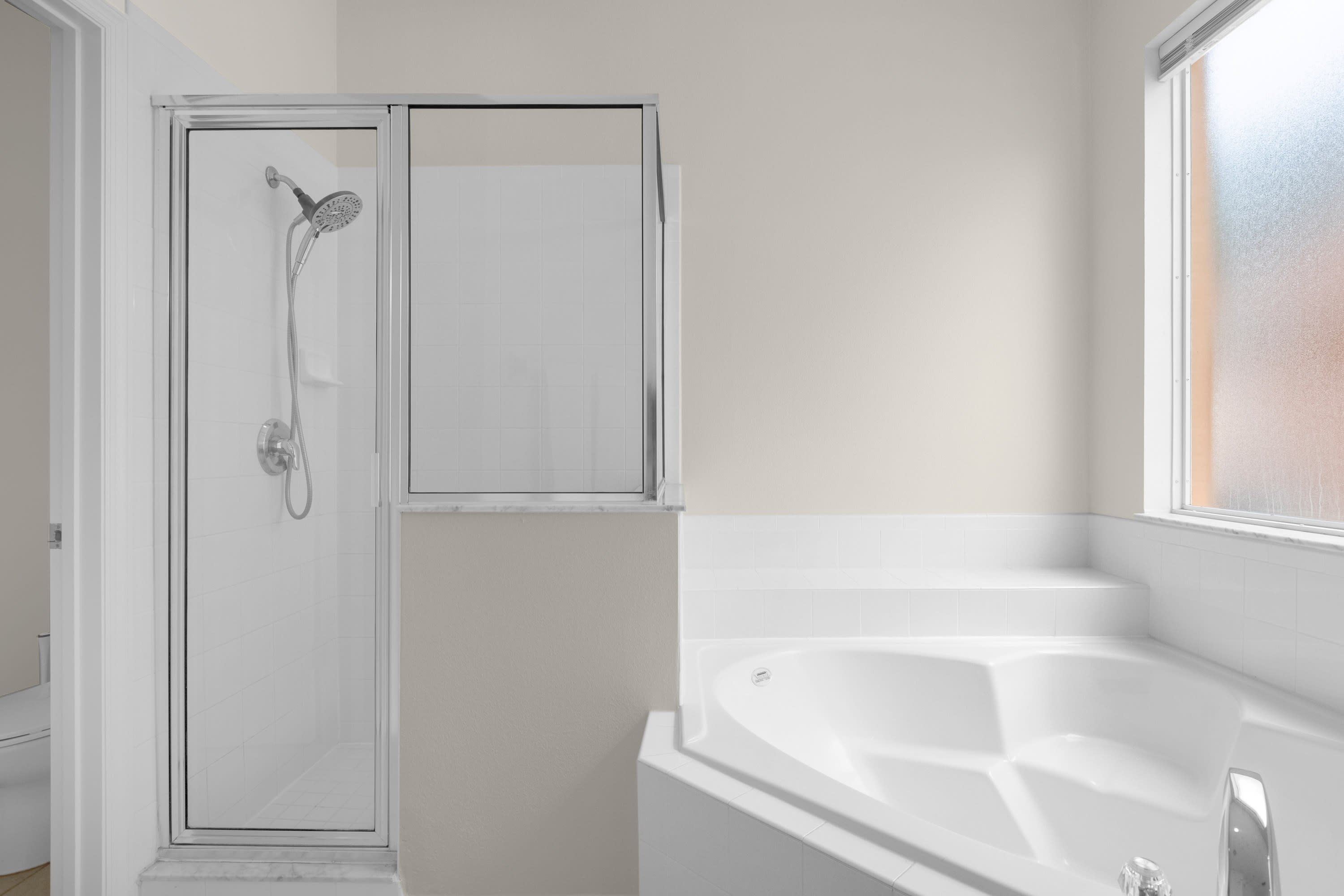 A close-up view of a bright bathroom highlights a white corner jetted tub with tiled steps and a modern glass-enclosed shower with a rainfall showerhead. The walls are painted a light beige, and a frosted window provides natural light.