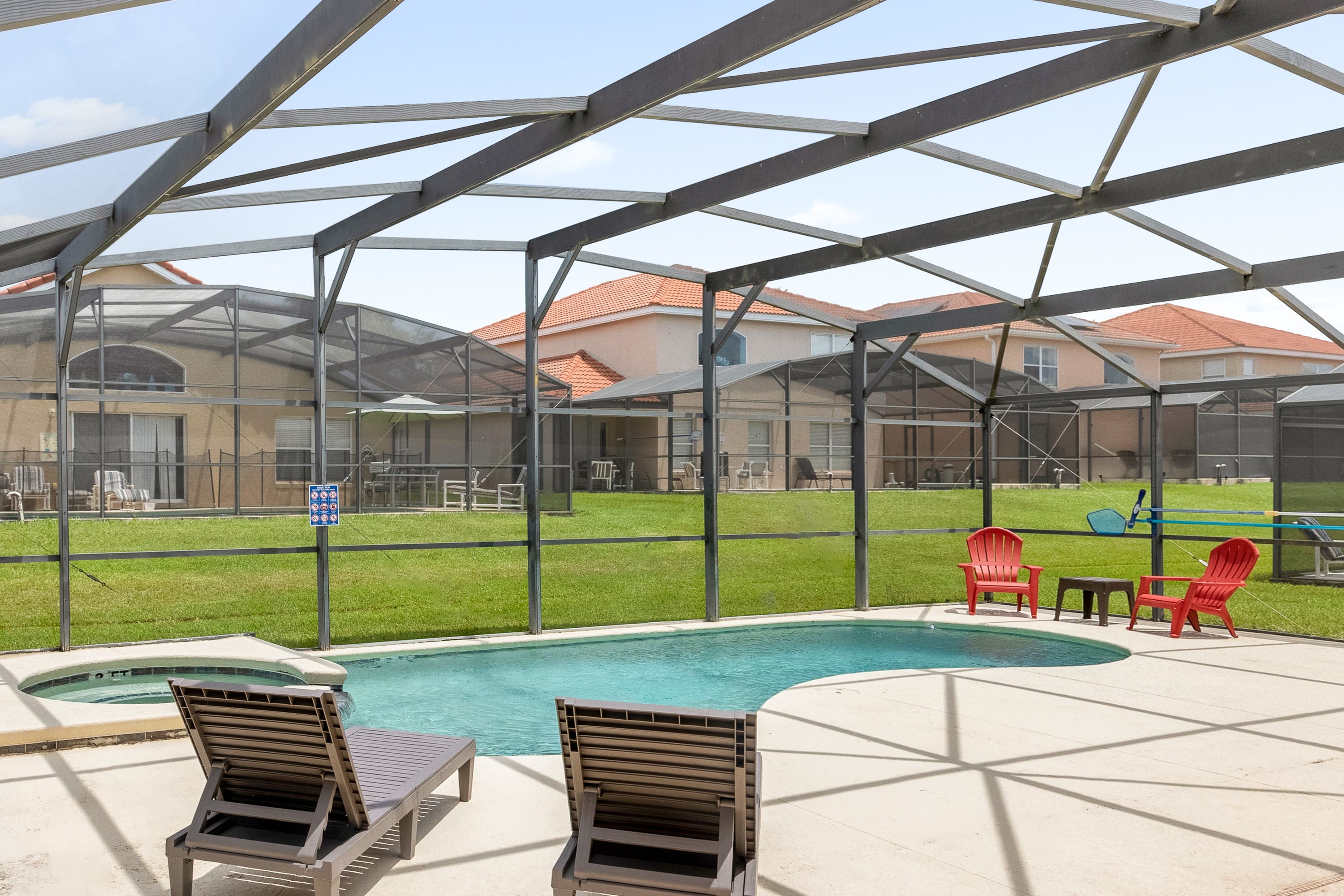A private, screened-in pool area features a small, kidney-shaped pool with an adjacent hot tub, surrounded by a light-colored concrete deck. Red Adirondack chairs and a small table are placed on the grass outside the screen, with other houses visible in the background.