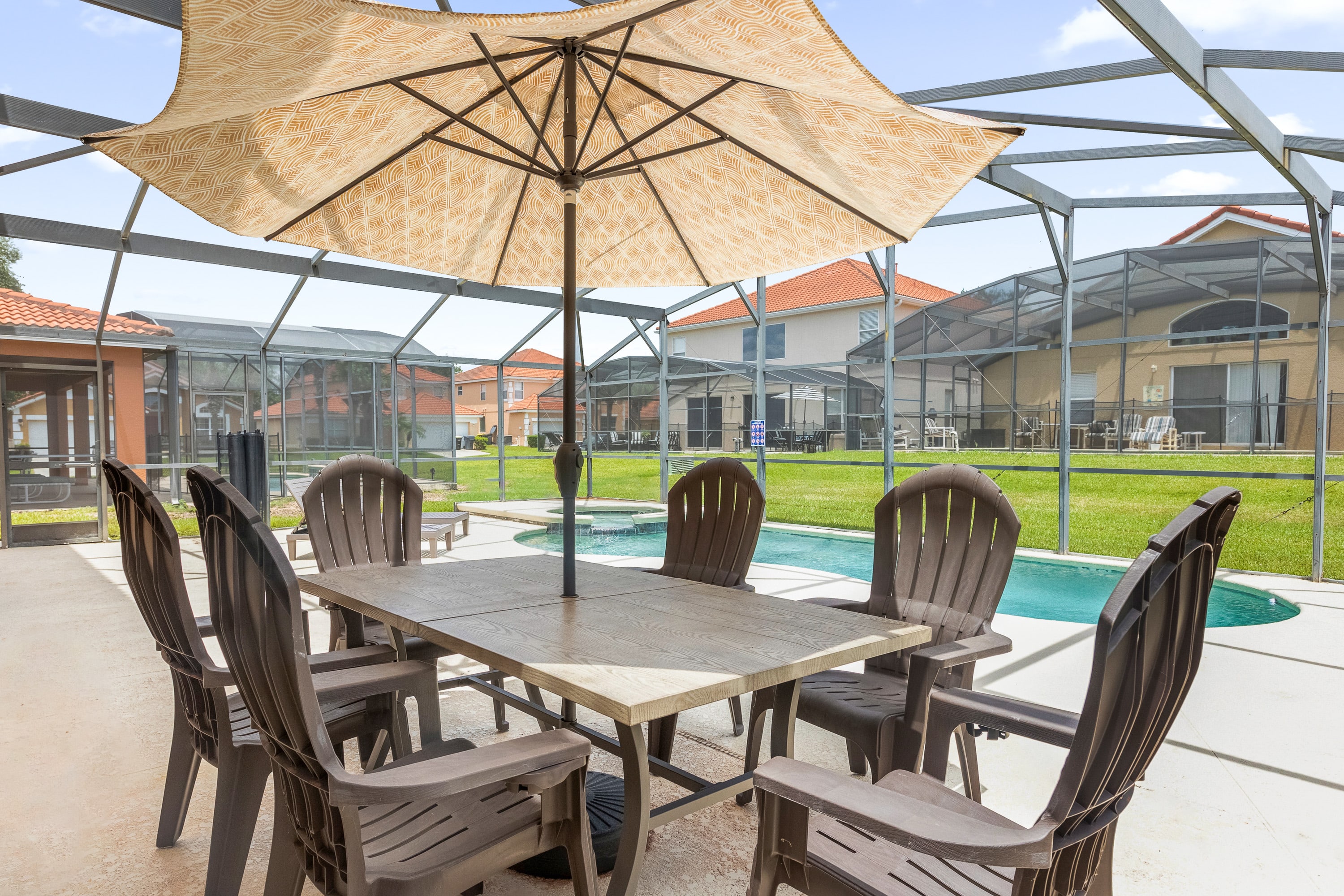 A close-up of the outdoor dining area on the screened-in patio features a large rectangular table with six brown chairs and a central umbrella providing shade. The private pool is visible in the background, surrounded by the light-colored concrete deck.