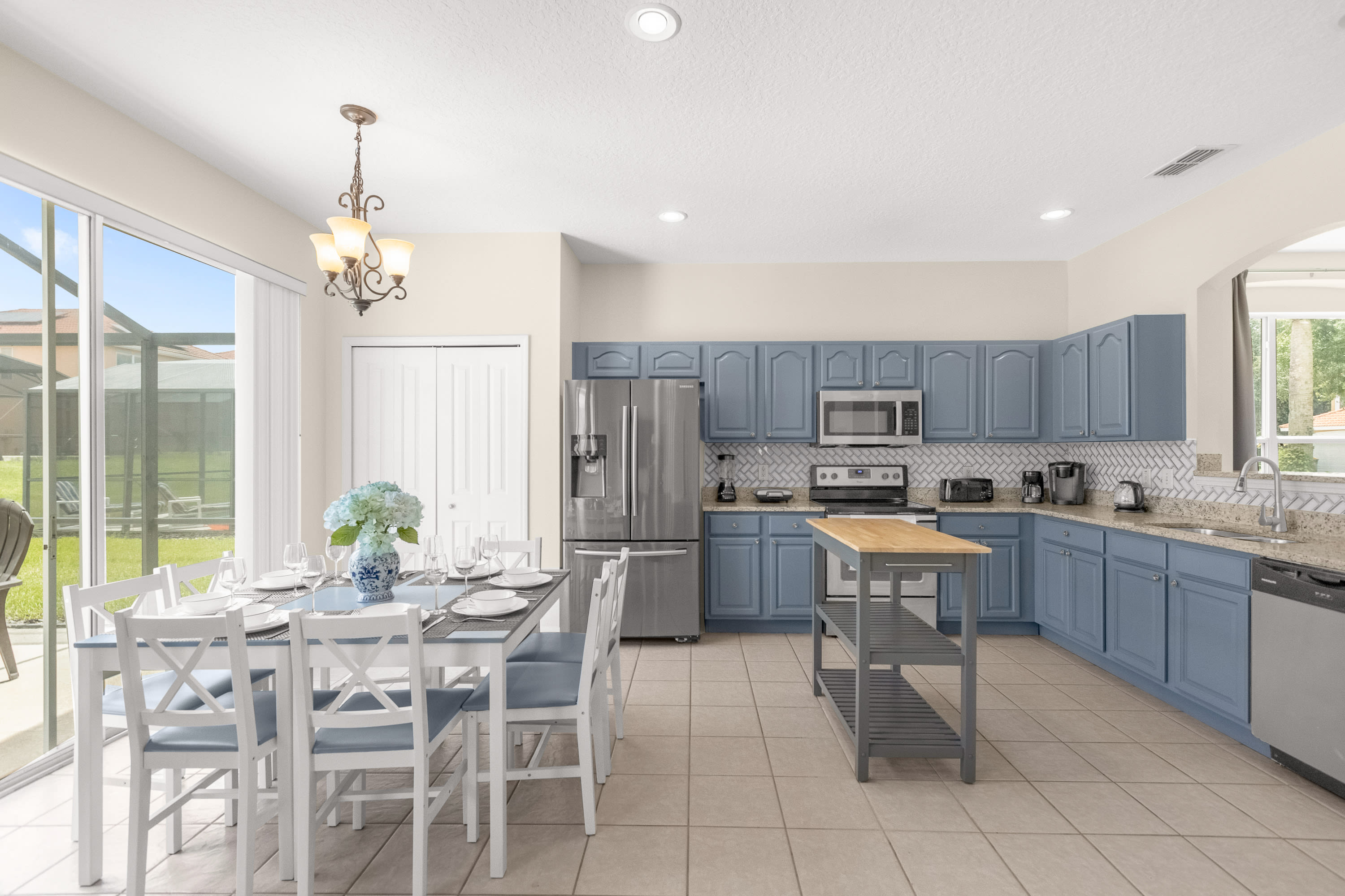 Spacious kitchen with light blue cabinets, granite countertops, white subway tile backsplash, and stainless steel appliances, including a refrigerator, microwave, and dishwasher. A portable wooden island and a dining table are in the foreground.