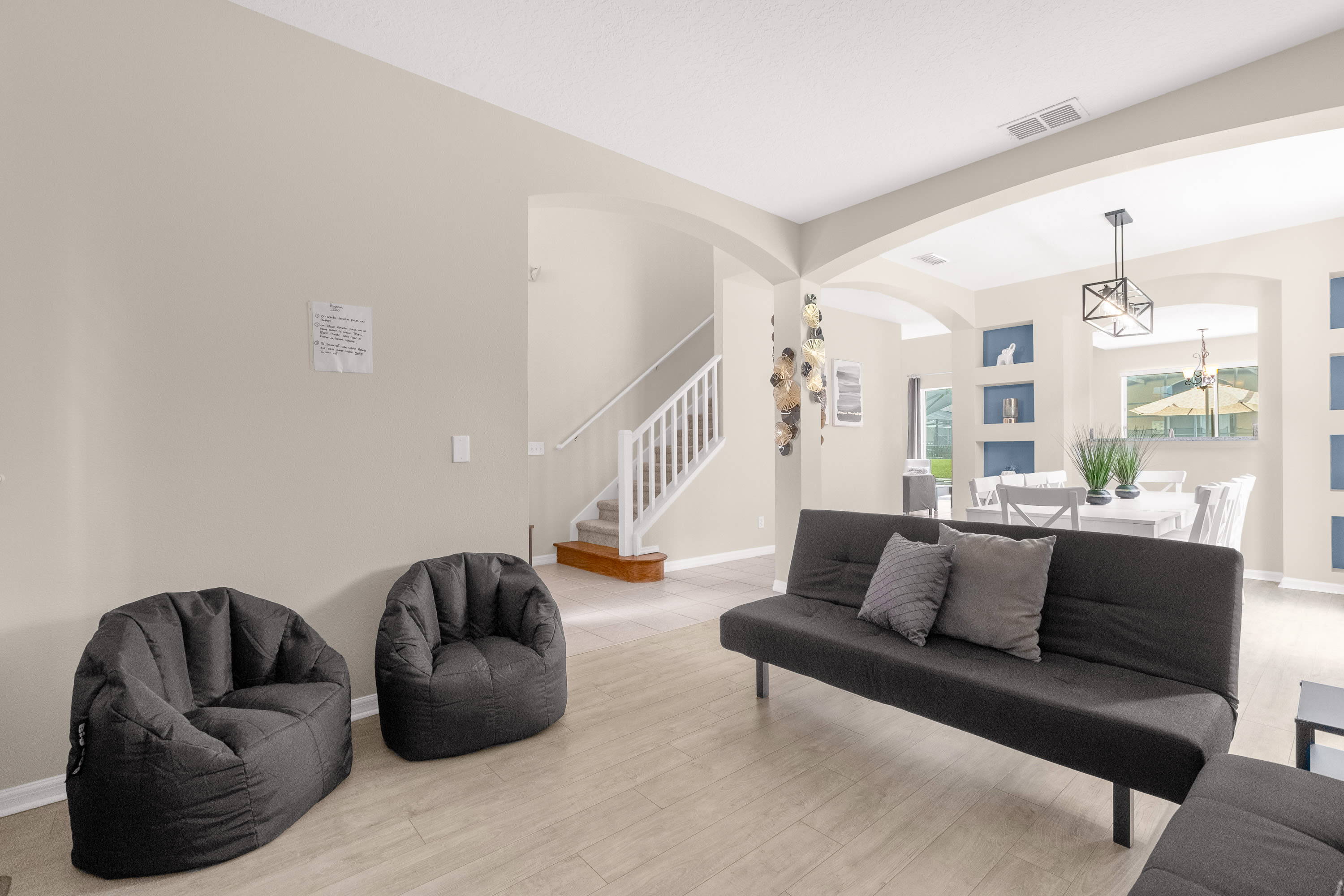 Open-concept living area with light wood-look flooring, two dark gray bean bag chairs, and a dark gray futon. A staircase and a dining area are visible in the background, featuring light-colored walls.