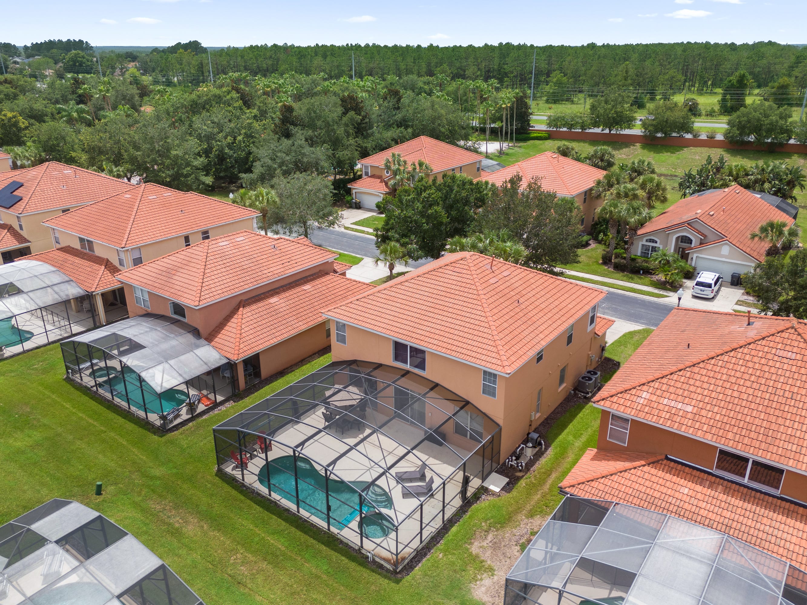 An aerial perspective showcasing a cluster of two-story vacation homes, each featuring a private screened-in pool and patio area in the backyard. The houses have warm-toned stucco exteriors and distinctive orange tile roofs, set amidst green lawns and tropical foliage.
