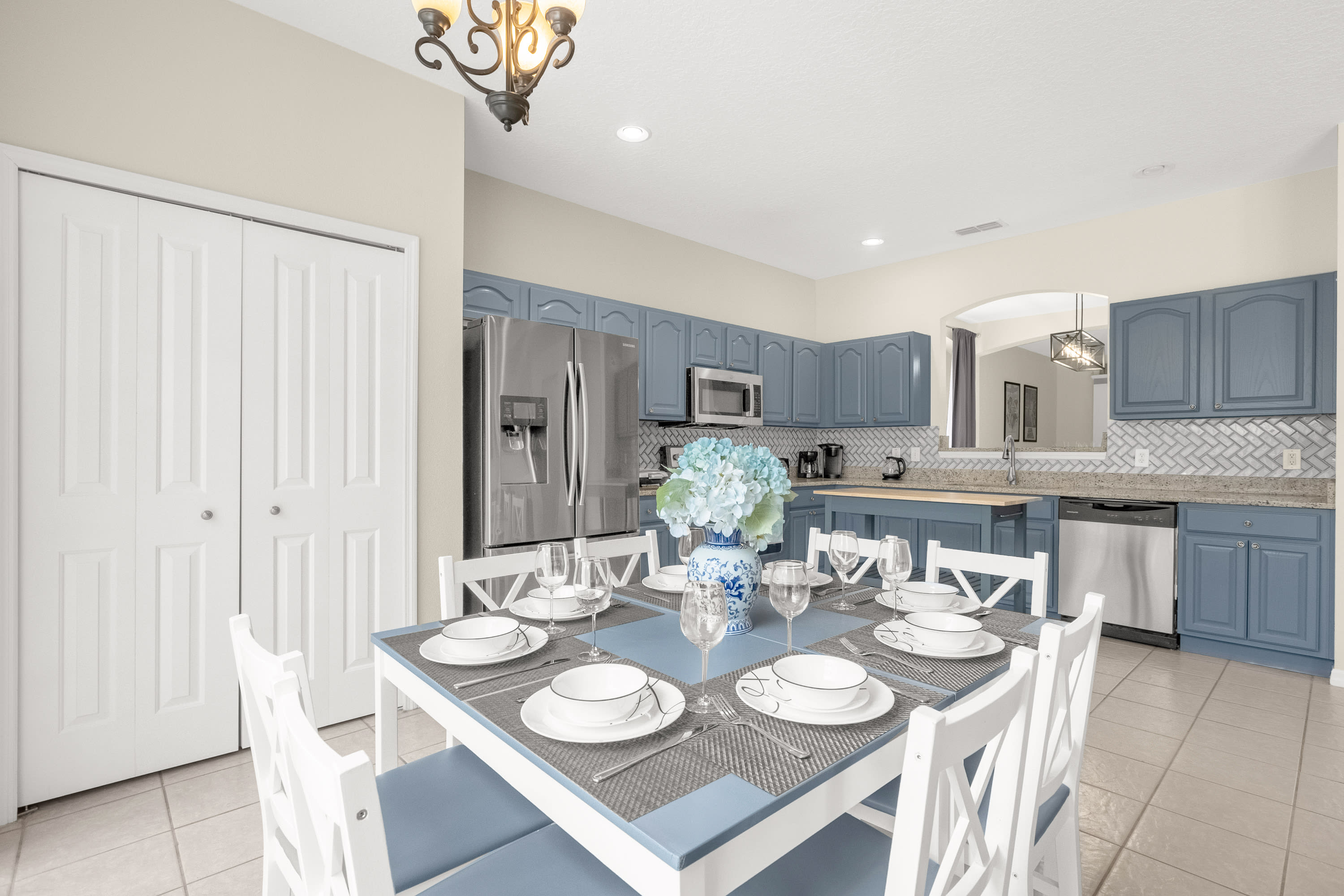 Dining area with a light blue and white dining table set for eight, featuring a blue floral centerpiece, positioned in front of a kitchen with blue cabinets, white subway tile backsplash, and stainless steel appliances.
