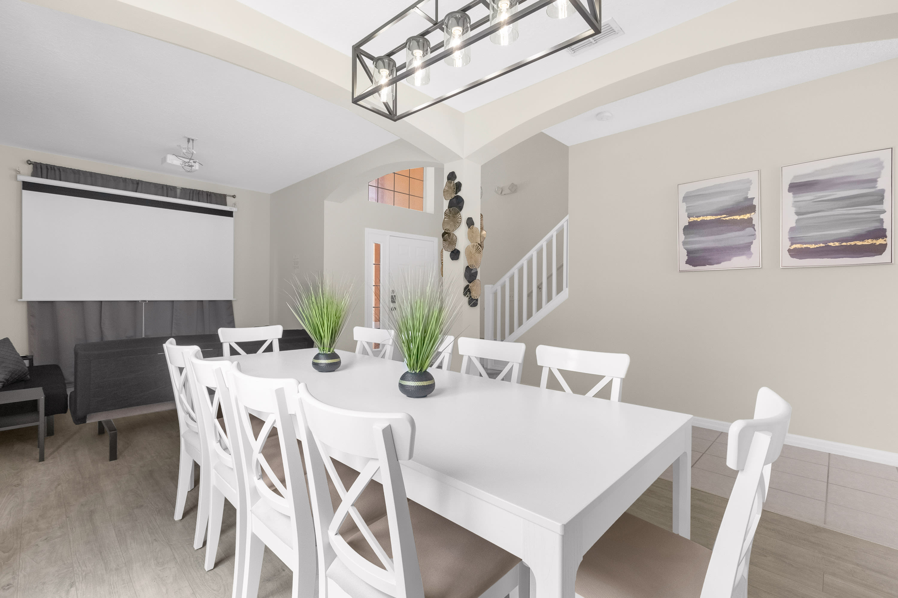 Spacious dining room with a long white dining table and eight white cross-back chairs, featuring a black linear chandelier, light wood-look flooring, and a view towards a media area with a projector screen and a staircase.