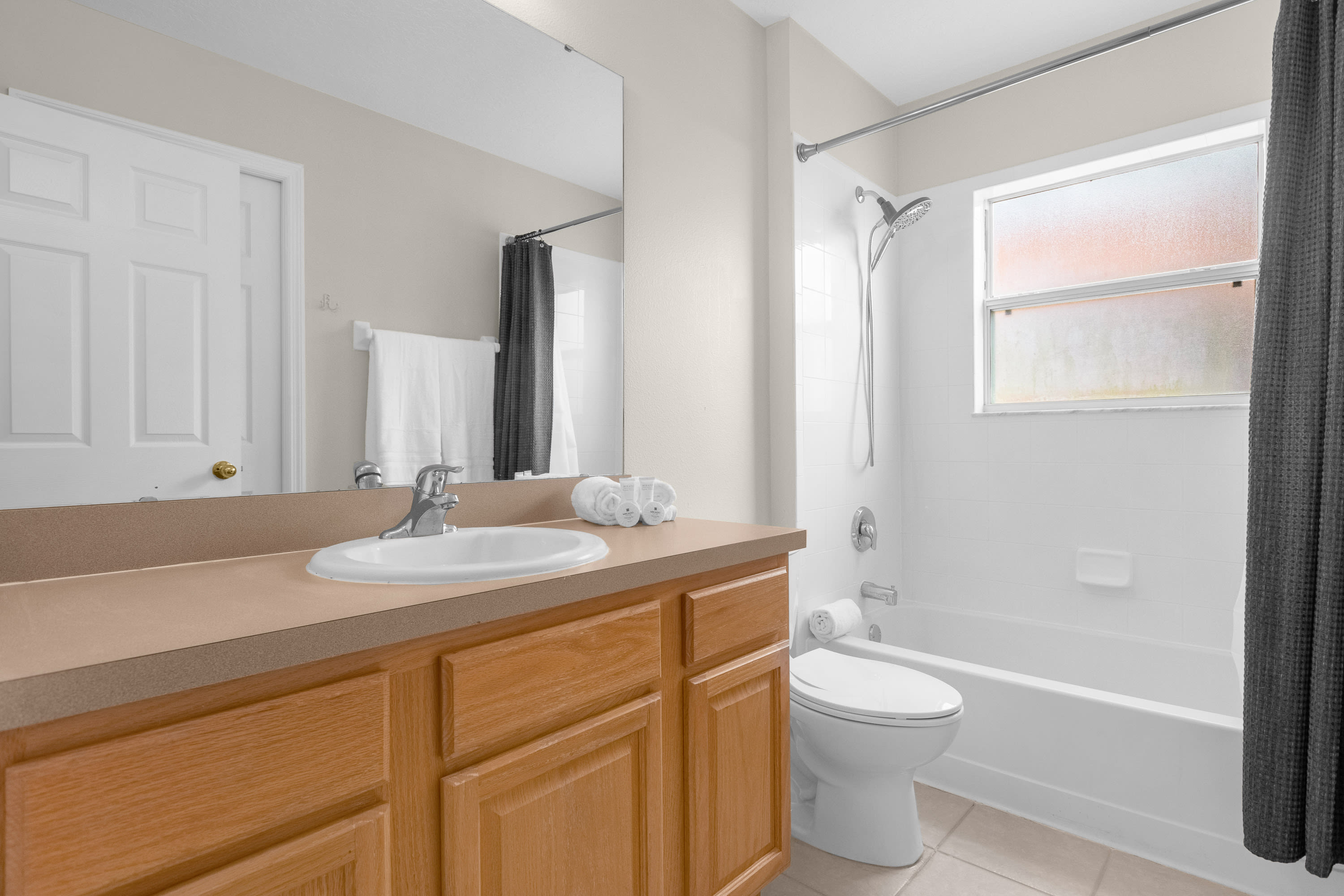 A bright bathroom featuring a light wood vanity with a single sink and a large mirror, alongside a white toilet and a shower-tub combo with white tiled walls and a window.