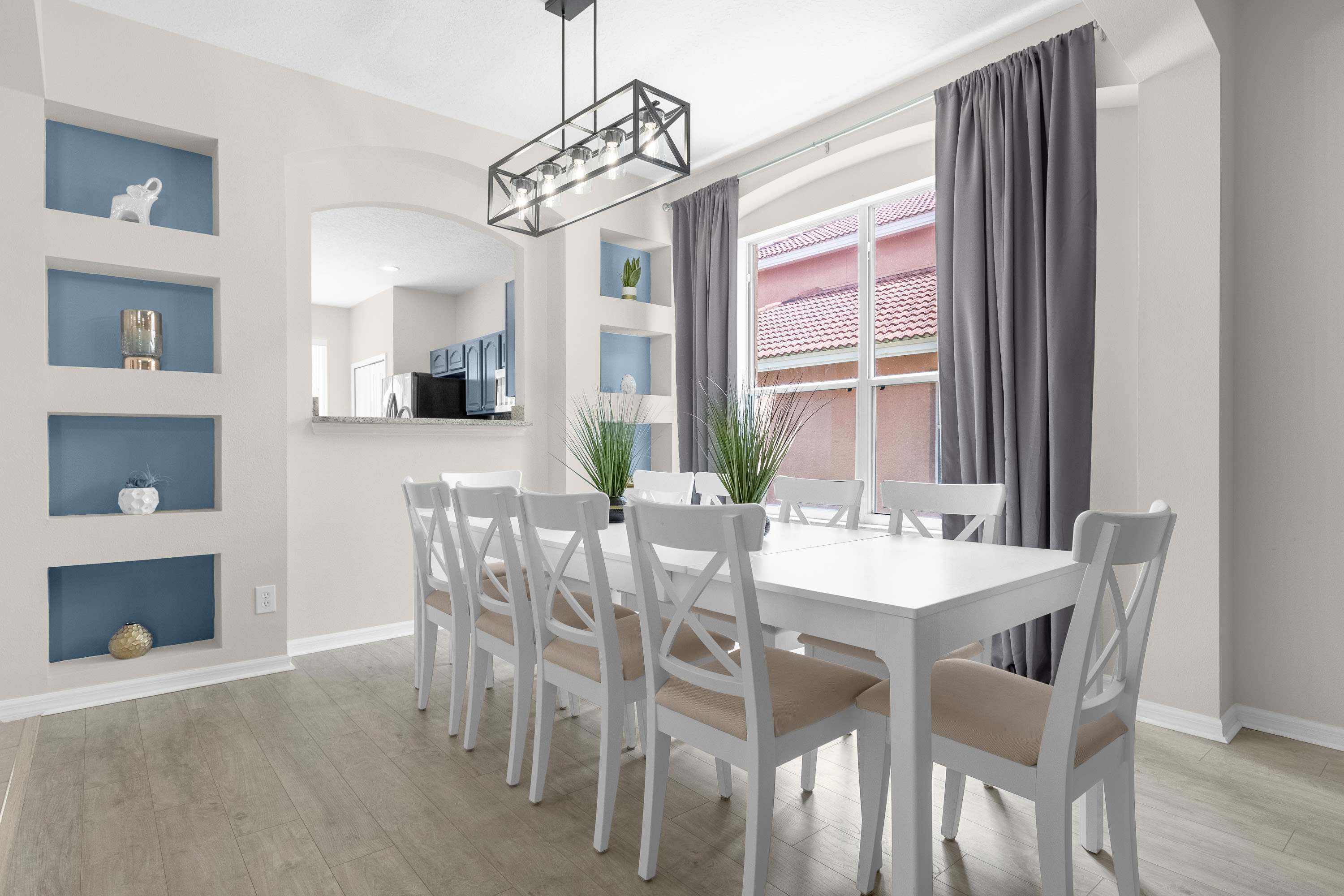 Elegant dining room with light wood-look flooring, a long white dining table with eight white cross-back chairs, and a black linear chandelier. It is complemented by built-in blue display shelves and a large window with gray curtains.