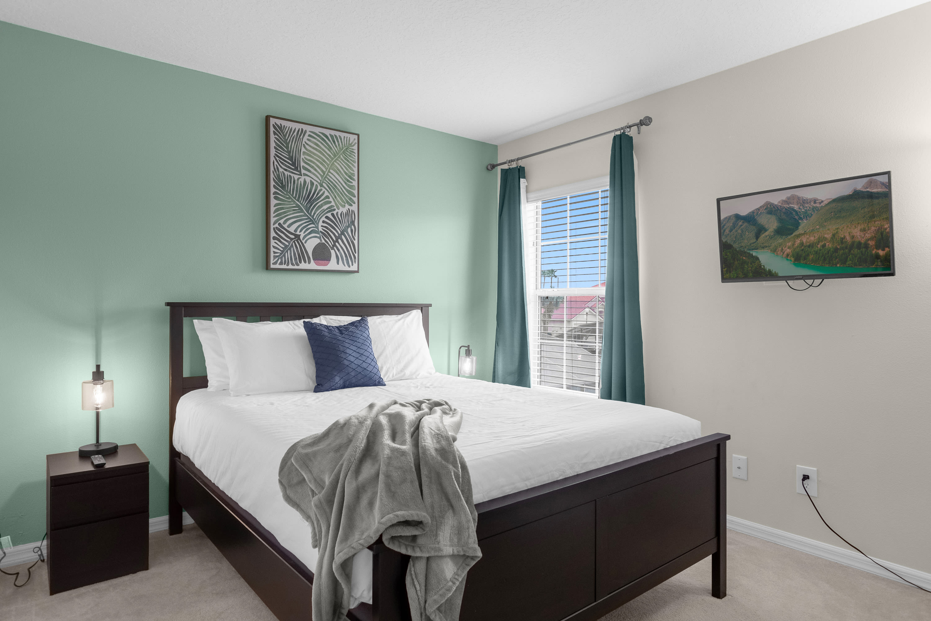 A cozy bedroom with a dark wood bed frame, white bedding, and a blue accent pillow, set against a two-tone wall with light green and beige, featuring a wall-mounted TV and a window with blinds.